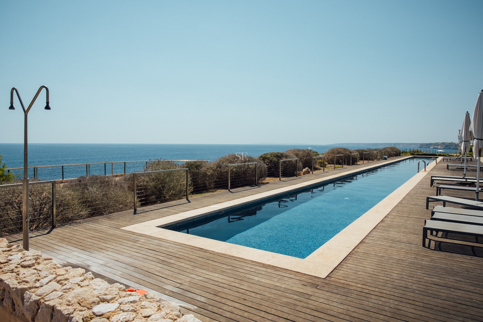 Long rectangular lap pool on a wooden deck overlooking the blue ocean with sun loungers and an outdoor shower.