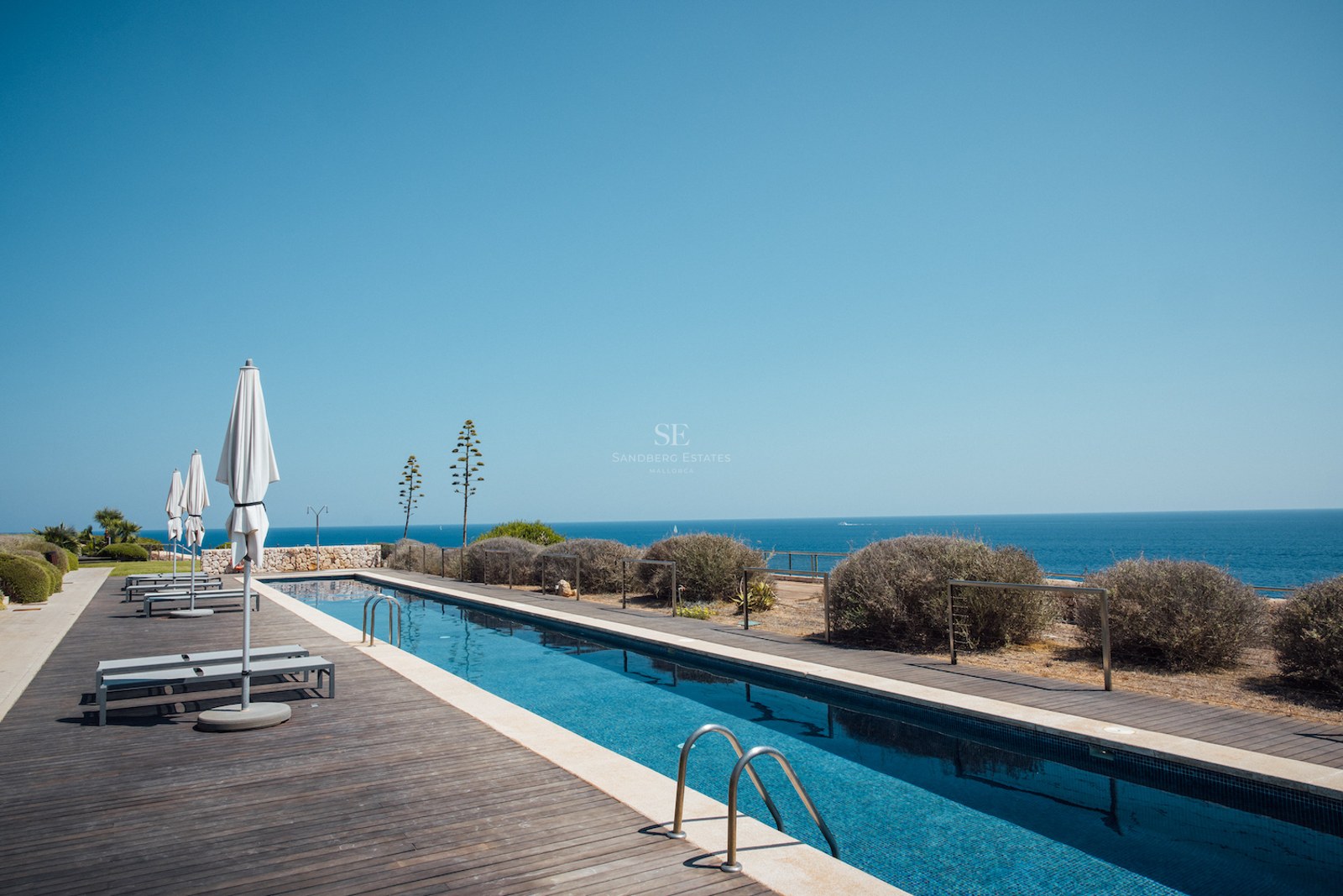 A long rectangular swimming pool with a dark wooden deck, white umbrellas, and an expansive view of the blue sea.