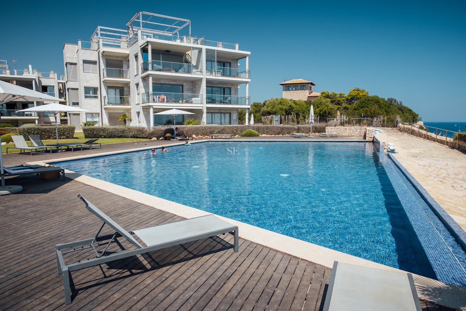 Large outdoor swimming pool with wooden deck, sun loungers, and modern apartment building under a clear blue sky.