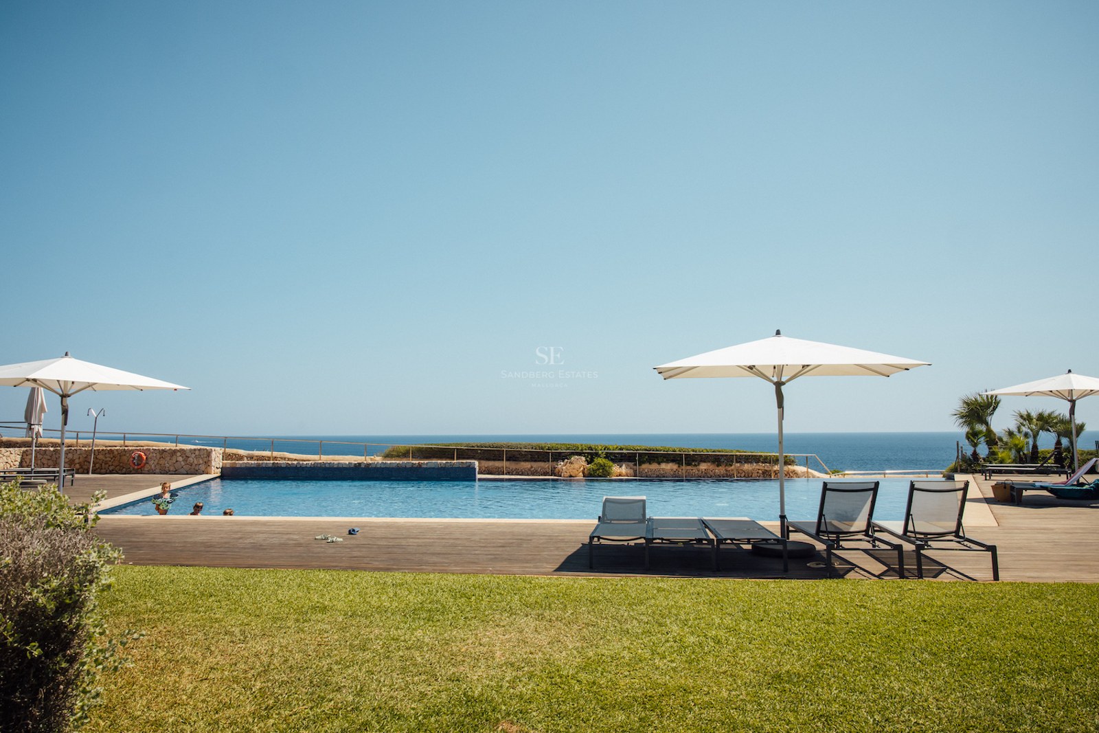 Large outdoor pool with wooden deck and sun loungers overlooking the sea under a clear blue sky.