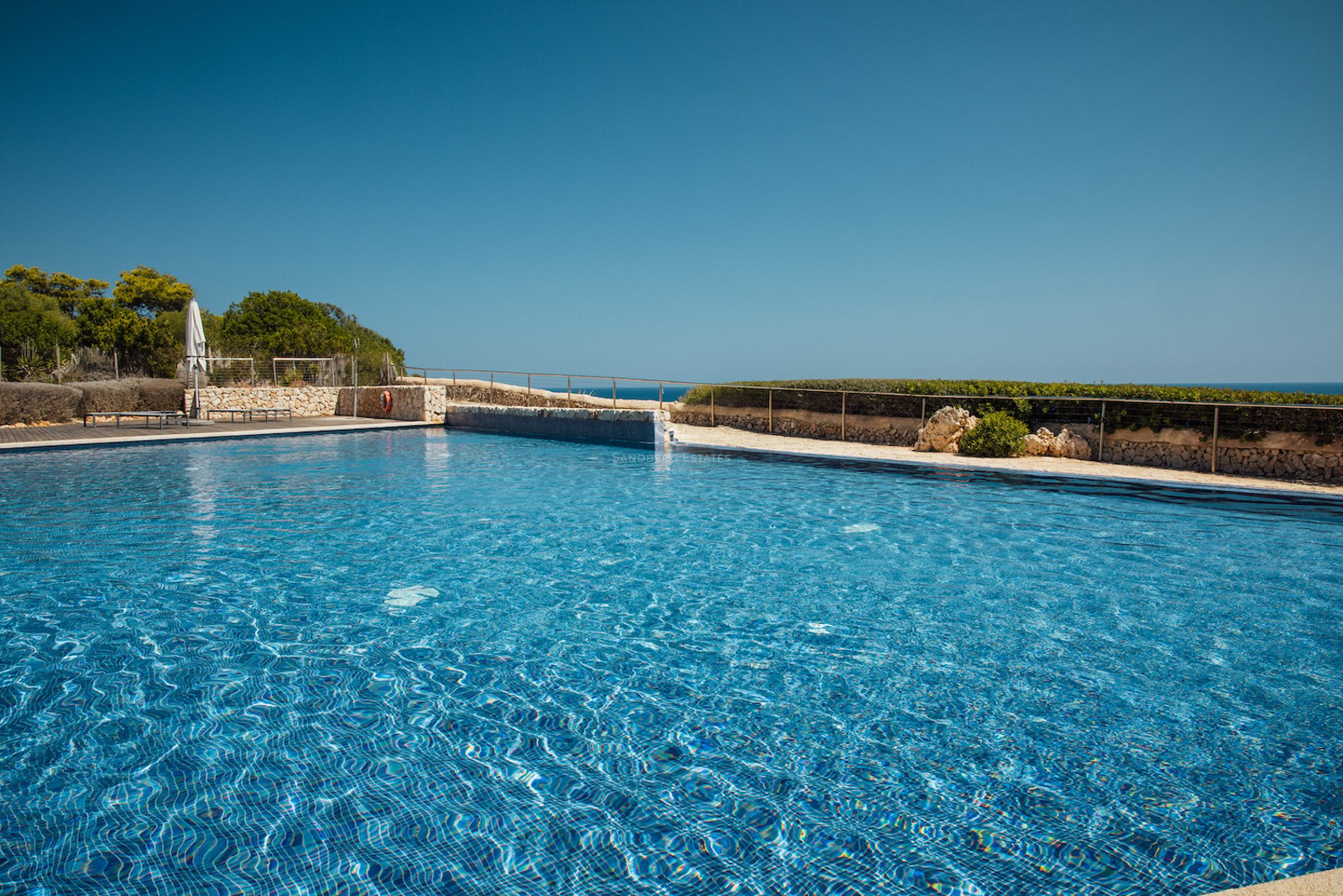 A large blue tiled swimming pool surrounded by natural stone walls and a metal railing under a clear blue sky.