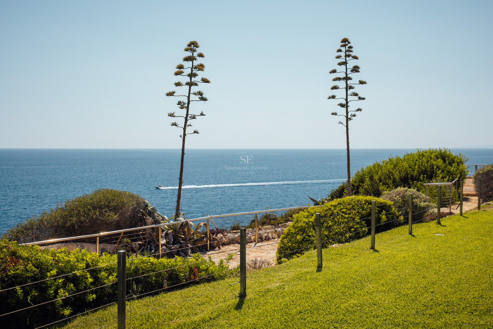 Coastal garden with a green lawn, two tall agave spikes, and an expansive view of the blue sea with a boat on the horizon.