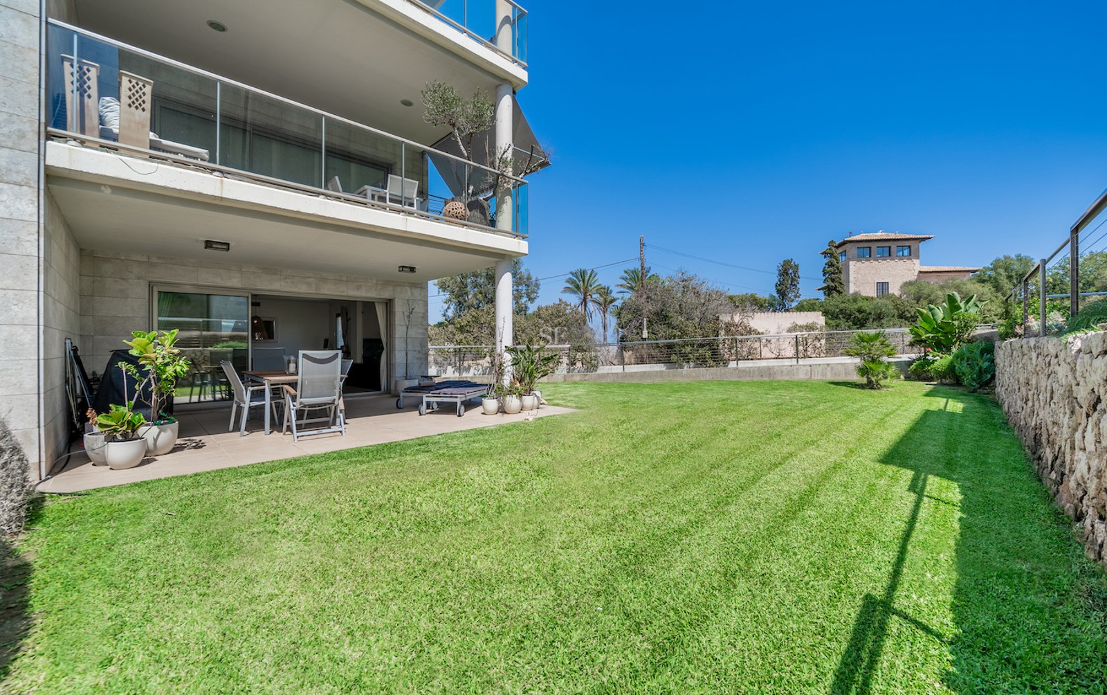 Covered ground floor terrace with outdoor dining area opening onto a manicured green lawn under a clear blue sky.