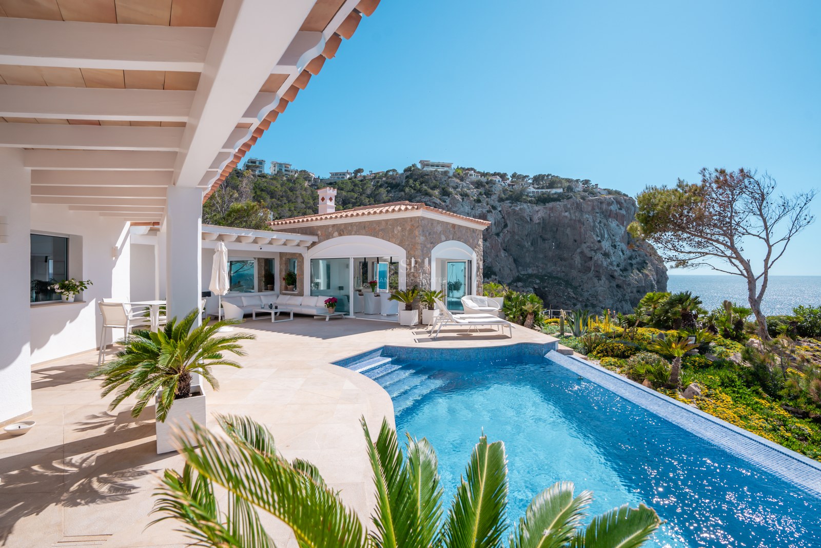 Curved infinity pool on a natural stone terrace overlooking coastal cliffs and the Mediterranean Sea.