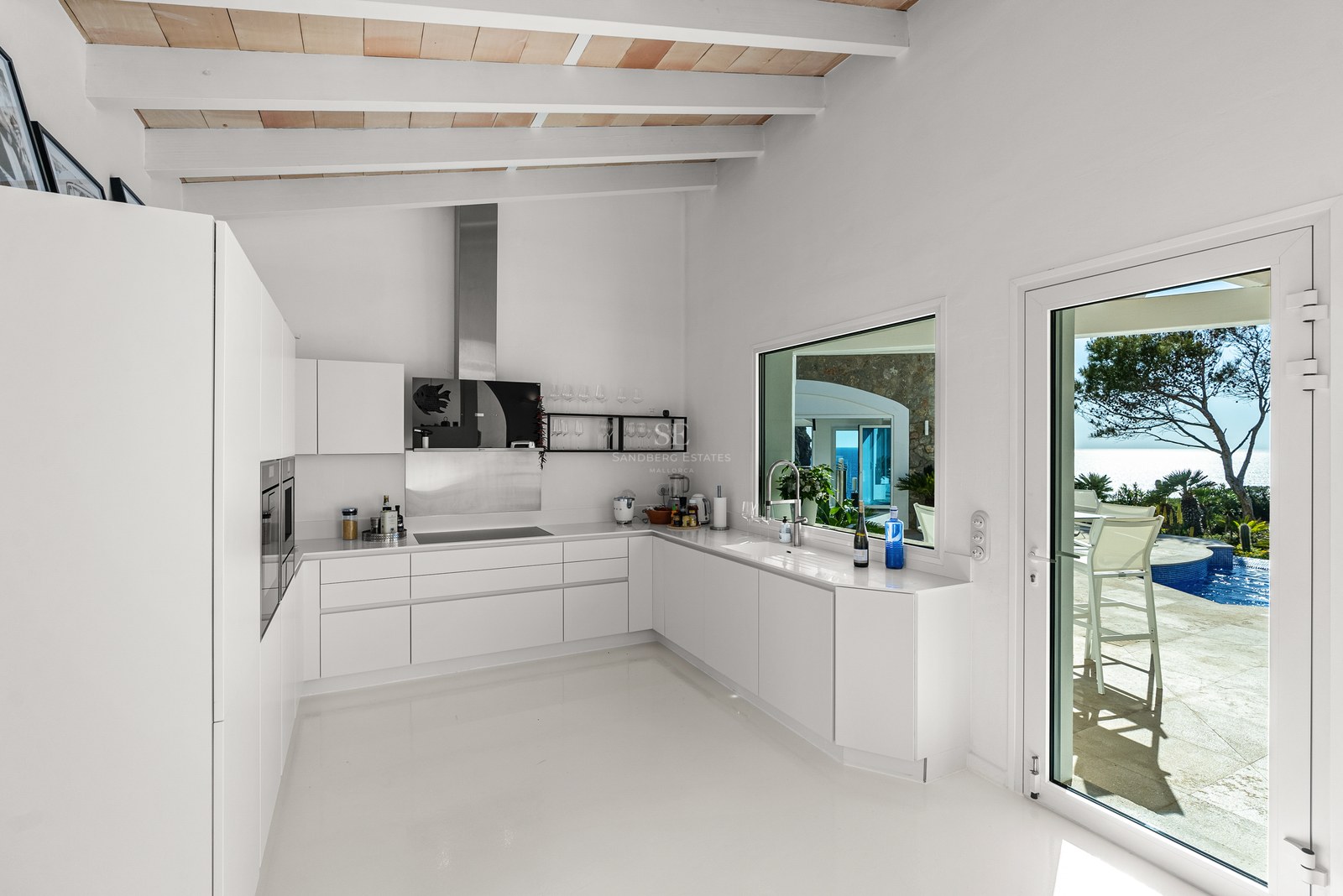 Sleek white minimalist kitchen featuring exposed wooden beams and glass door leading to a pool terrace with sea views.