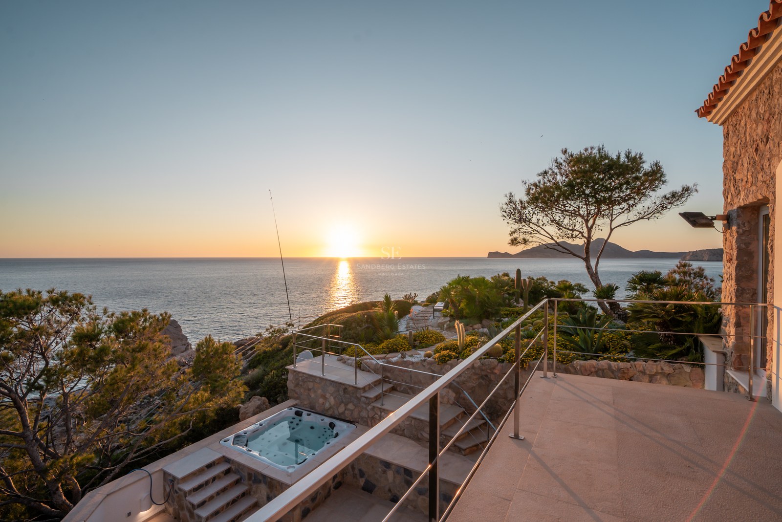 A multi-level stone terrace with a built-in jacuzzi overlooks the Mediterranean sea during a golden sunset.