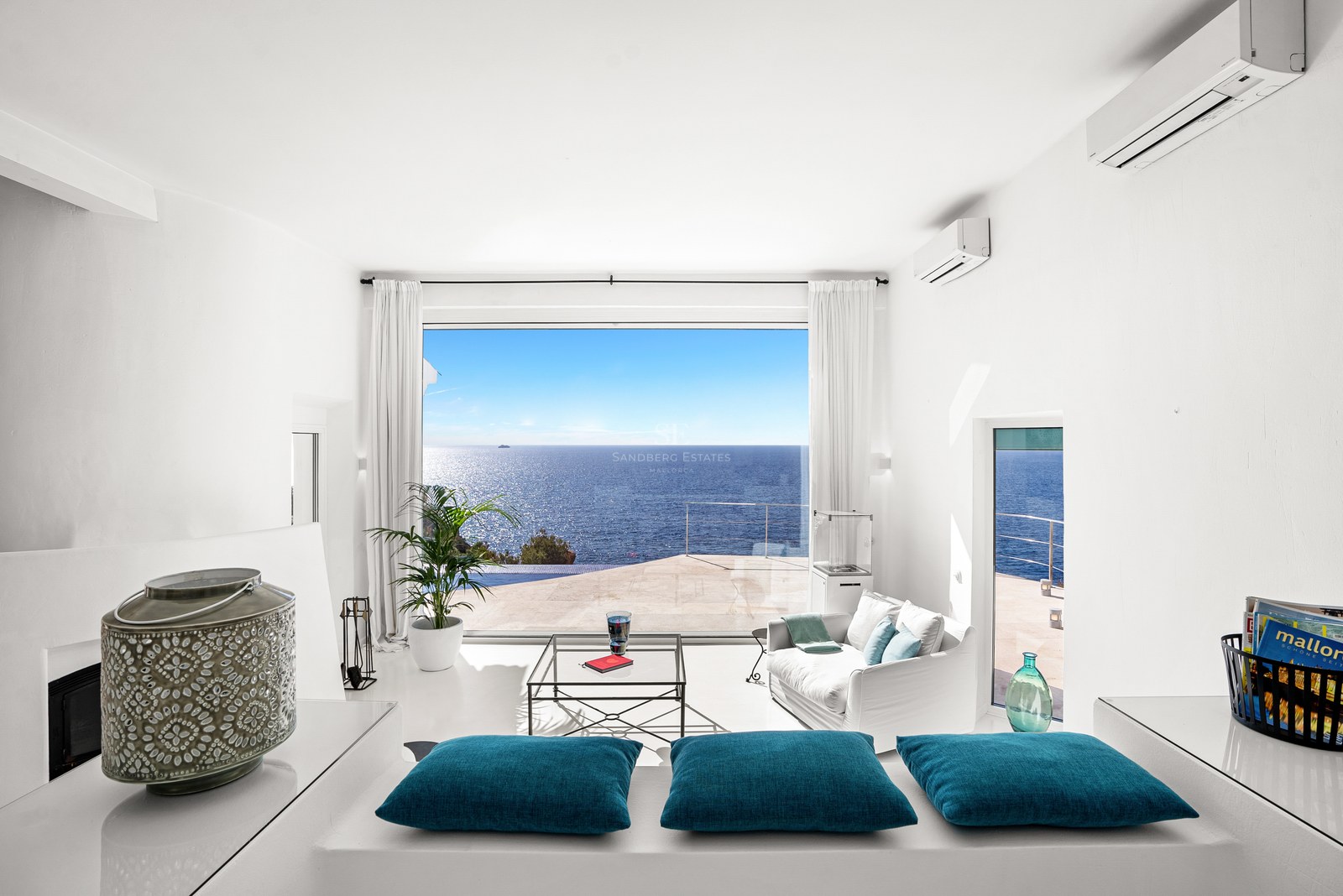Bright white minimalist living room with floor-to-ceiling windows overlooking a Mediterranean terrace and the blue sea.