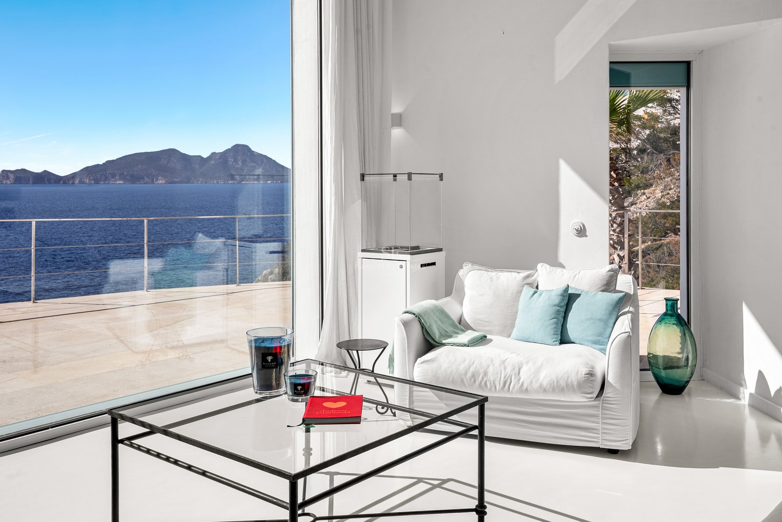 Interior of a white minimalist living room featuring a glass coffee table and armchair overlooking the sea.