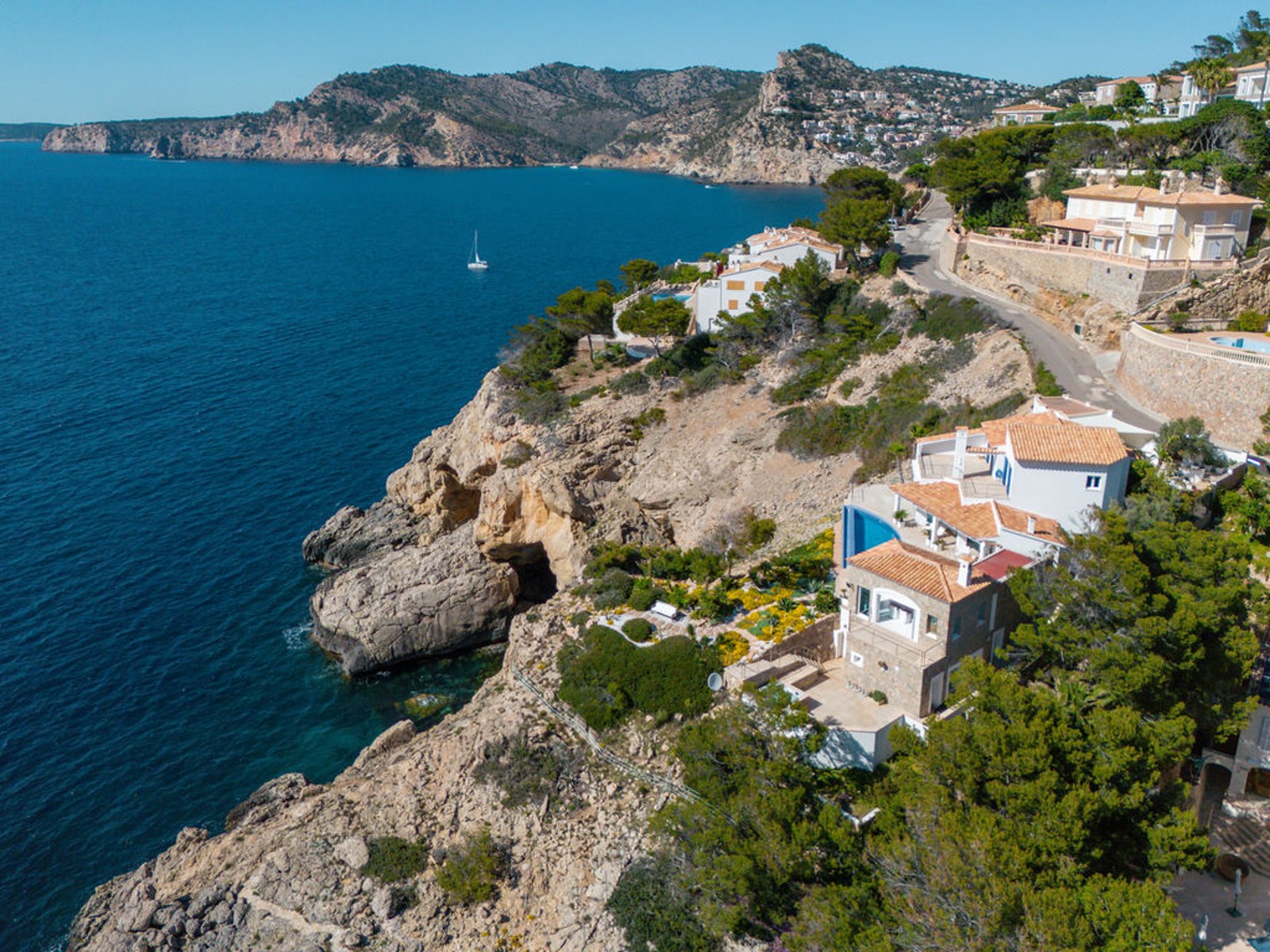 Drone view of a Mediterranean villa perched on a cliff above blue sea with a rocky coastline.