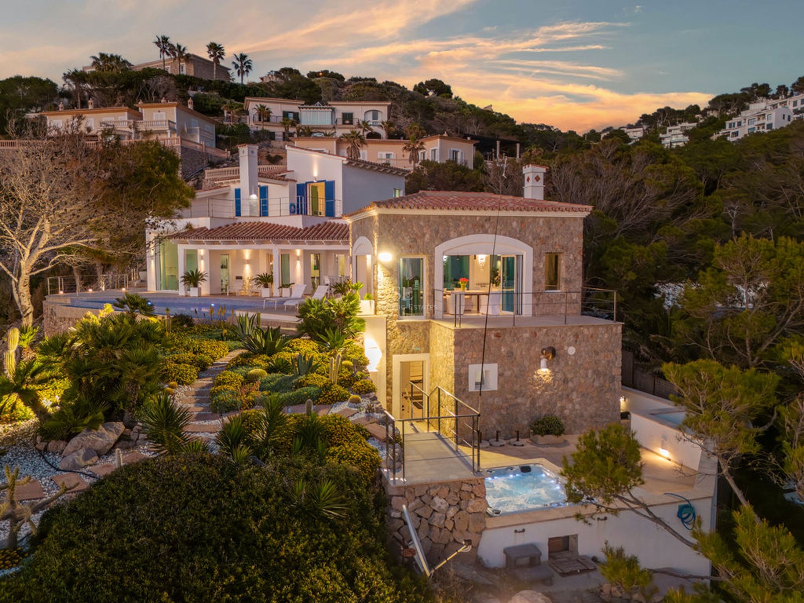 Exterior dusk shot of a multi-level stone villa featuring an illuminated pool, jacuzzi, and lush Mediterranean garden.