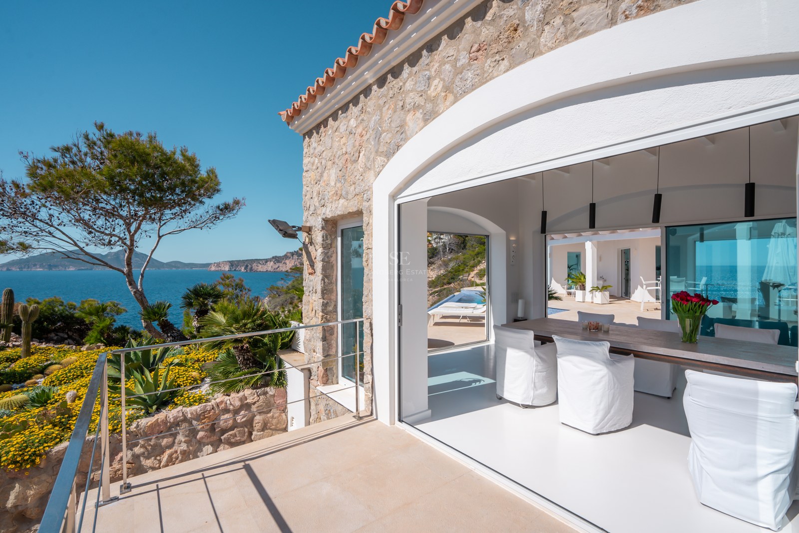 Modern dining room with wood table and white chairs opening to a sunny terrace with panoramic Mediterranean sea views.