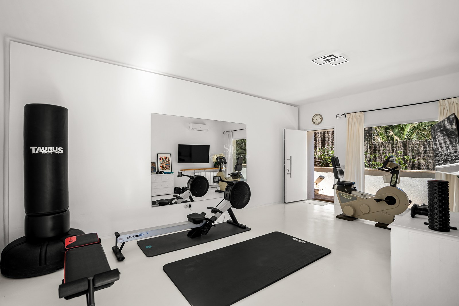 Modern white home gym with rowing machine, boxing bag, and exercise bike, featuring a large mirror and terrace access.