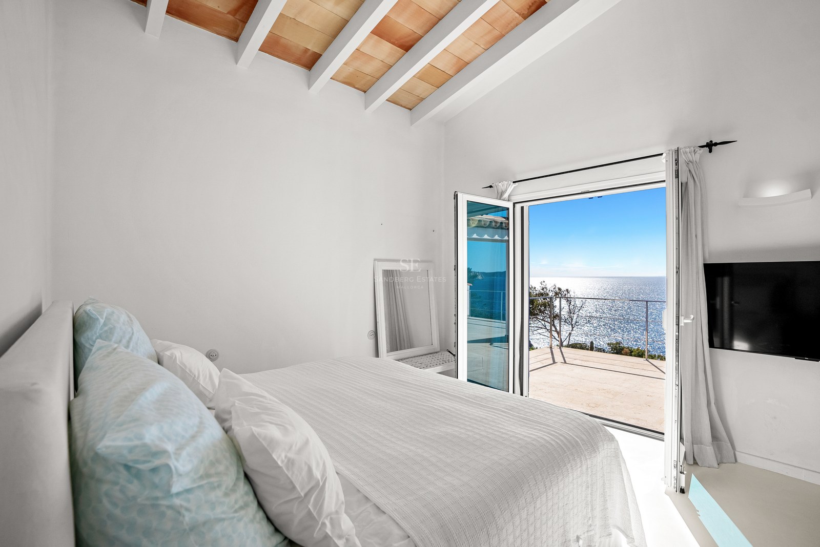 Bright master bedroom featuring white exposed ceiling beams and glass doors opening to a panoramic Mediterranean sea view.