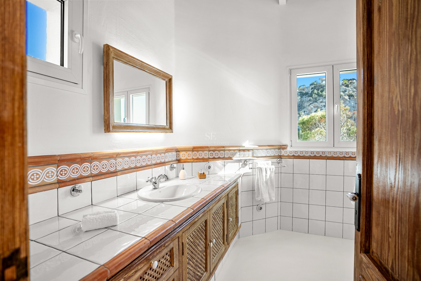 Bright bathroom featuring white tiles, terracotta accents, a wooden lattice vanity, and mountain views through windows.
