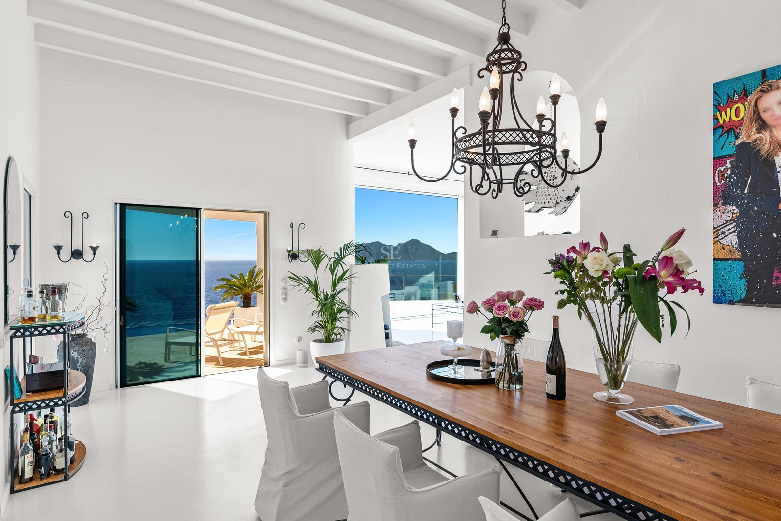 Modern dining room with a large wooden table, white beamed ceiling, and floor-to-ceiling windows overlooking the Mediterranean.