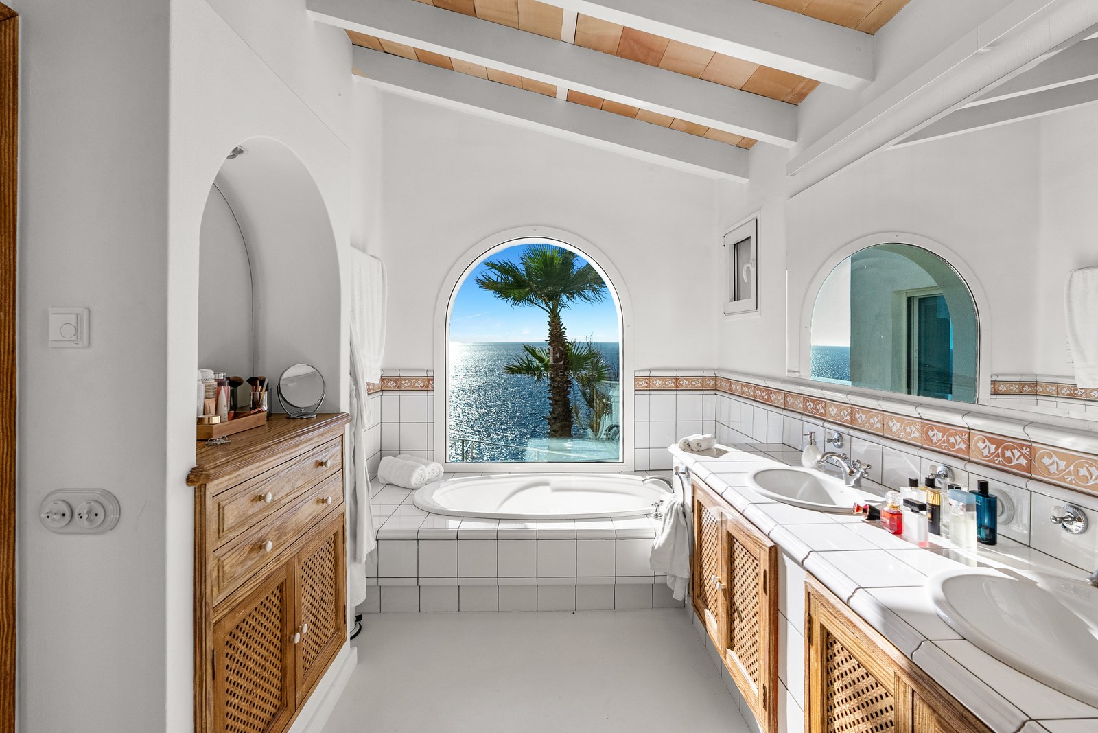 Bright white bathroom featuring a bathtub set before an arched window with Mediterranean sea views and beamed ceiling.