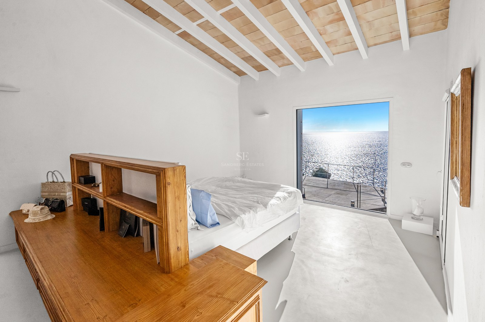 Minimalist white bedroom with custom wooden furniture and a large window overlooking the sparkling Mediterranean sea.