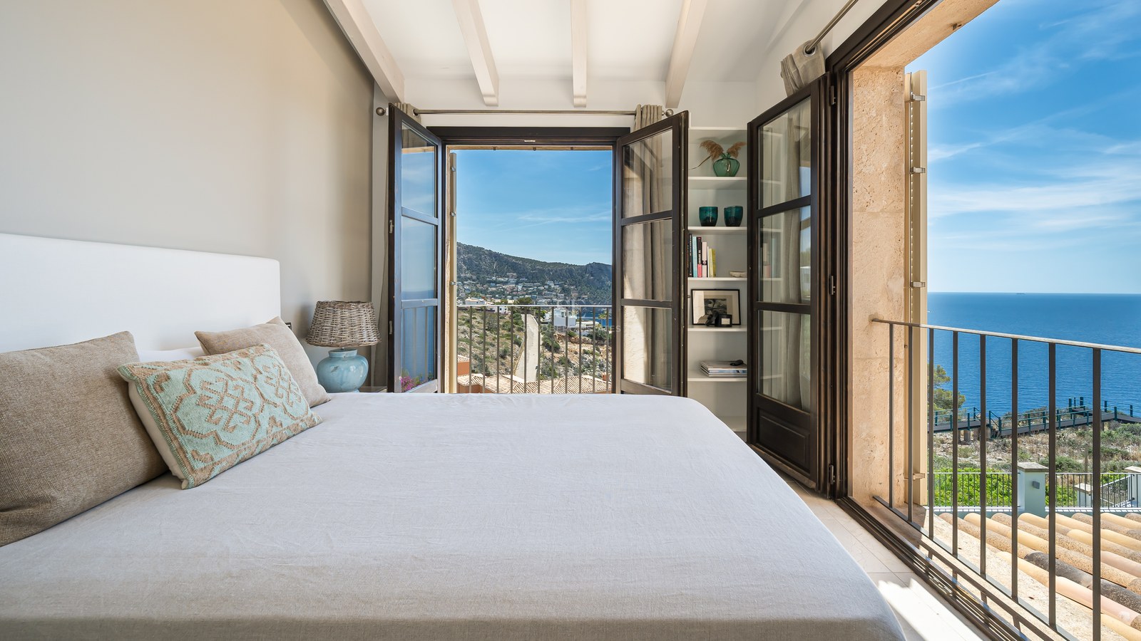 Chambre de luxe avec poutres blanches et portes-fenêtres ouvertes sur le littoral méditerranéen.