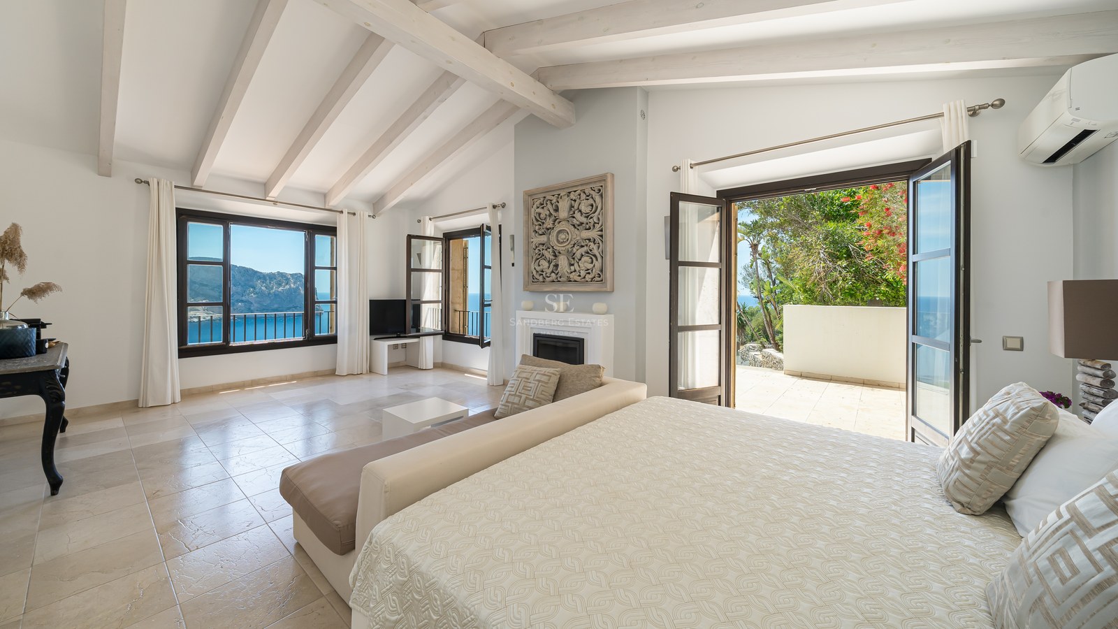 Chambre de maître lumineuse avec poutres blanches, sol en pierre et terrasse privée donnant sur la mer.