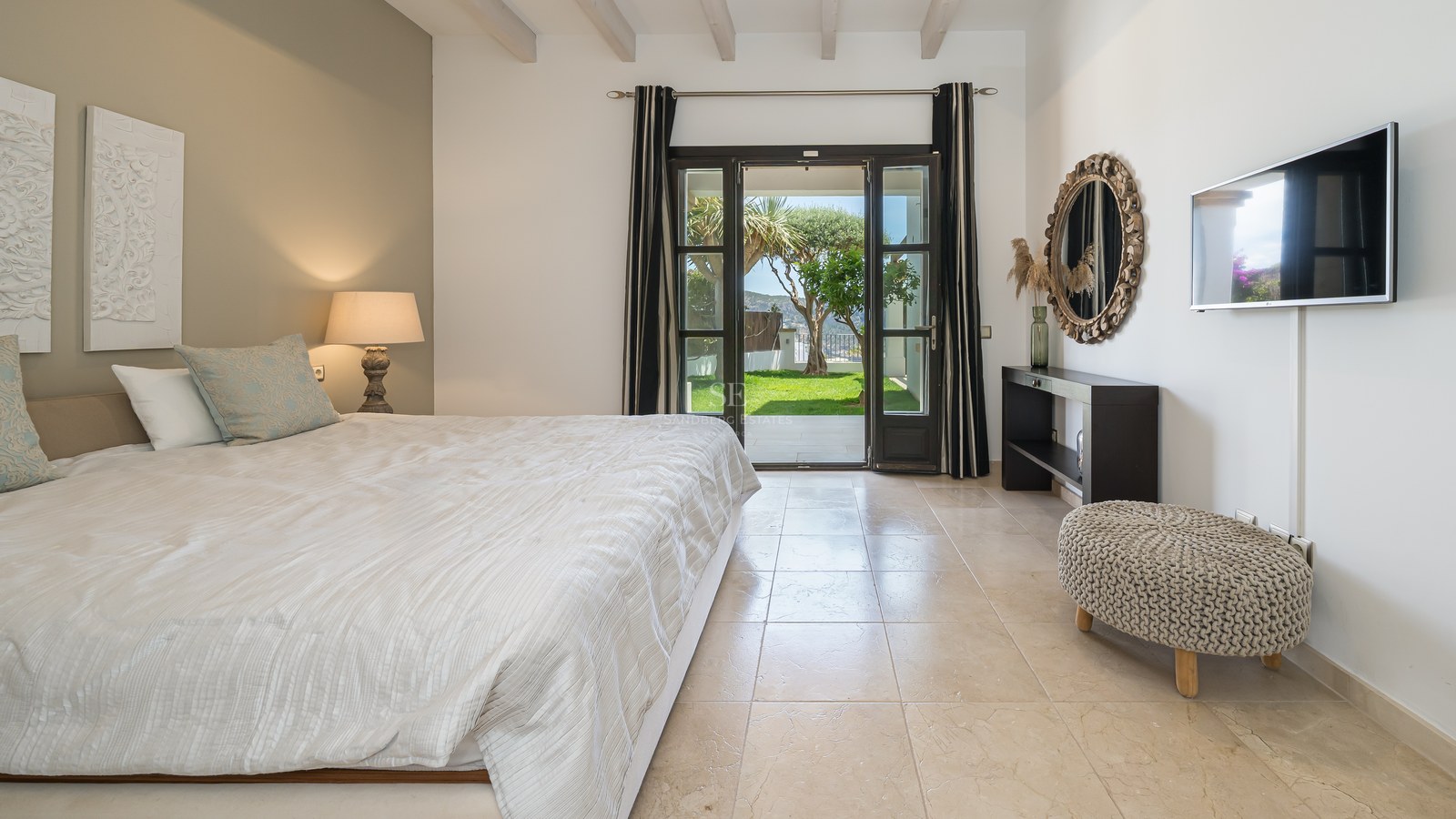 Chambre spacieuse avec sol en pierre naturelle, poutres blanches et portes vitrées ouvrant sur un jardin verdoyant.