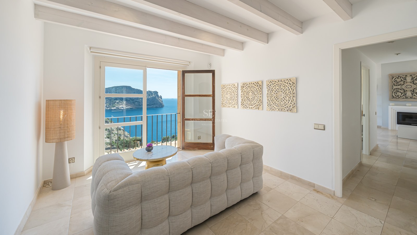 Salon lumineux avec poutres blanches, sol en pierre naturelle et balcon donnant sur la mer bleue et les falaises.