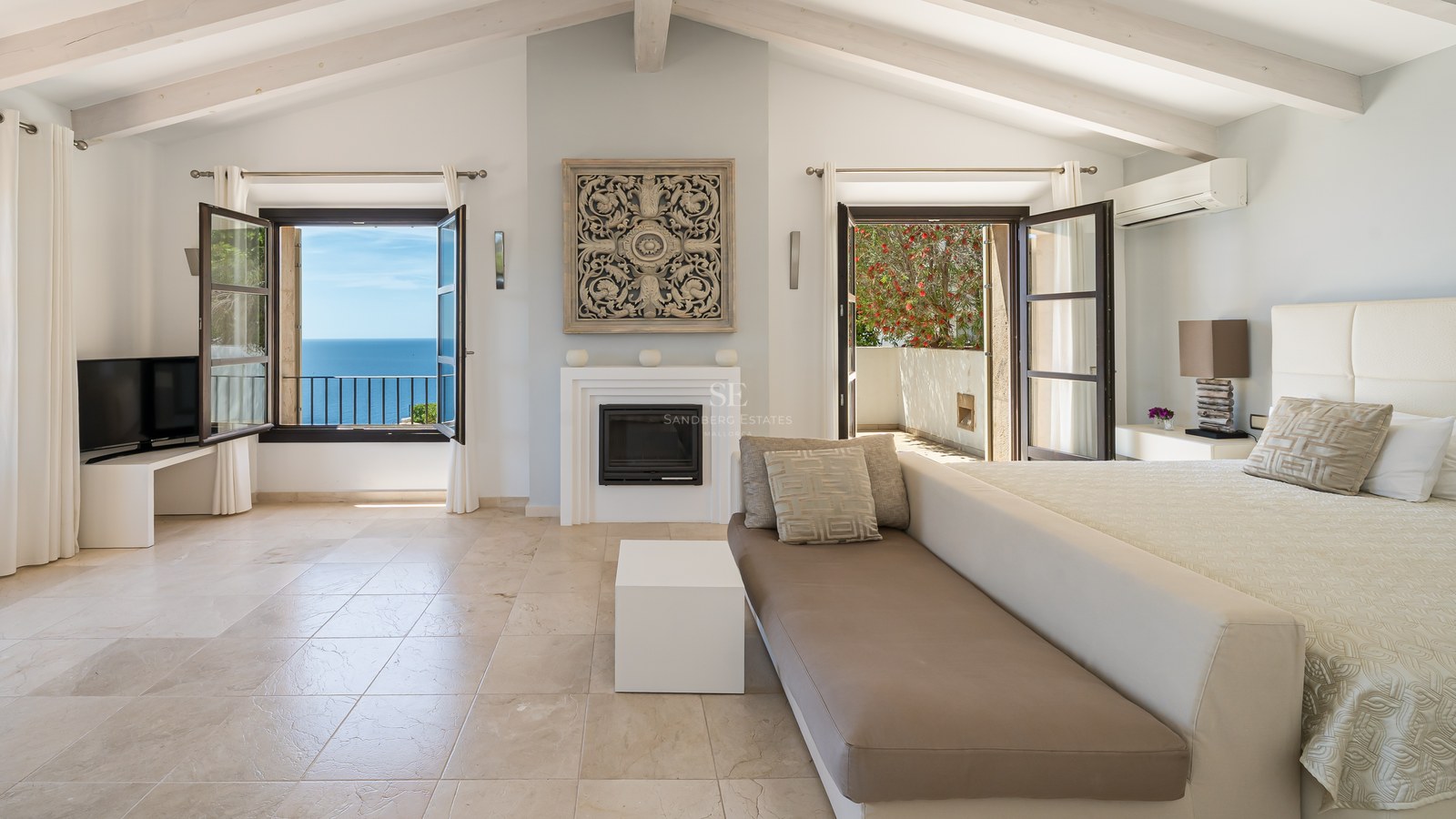 Grande chambre avec plafonds à poutres blanches, cheminée, sol en pierre et fenêtres ouvertes sur la mer.