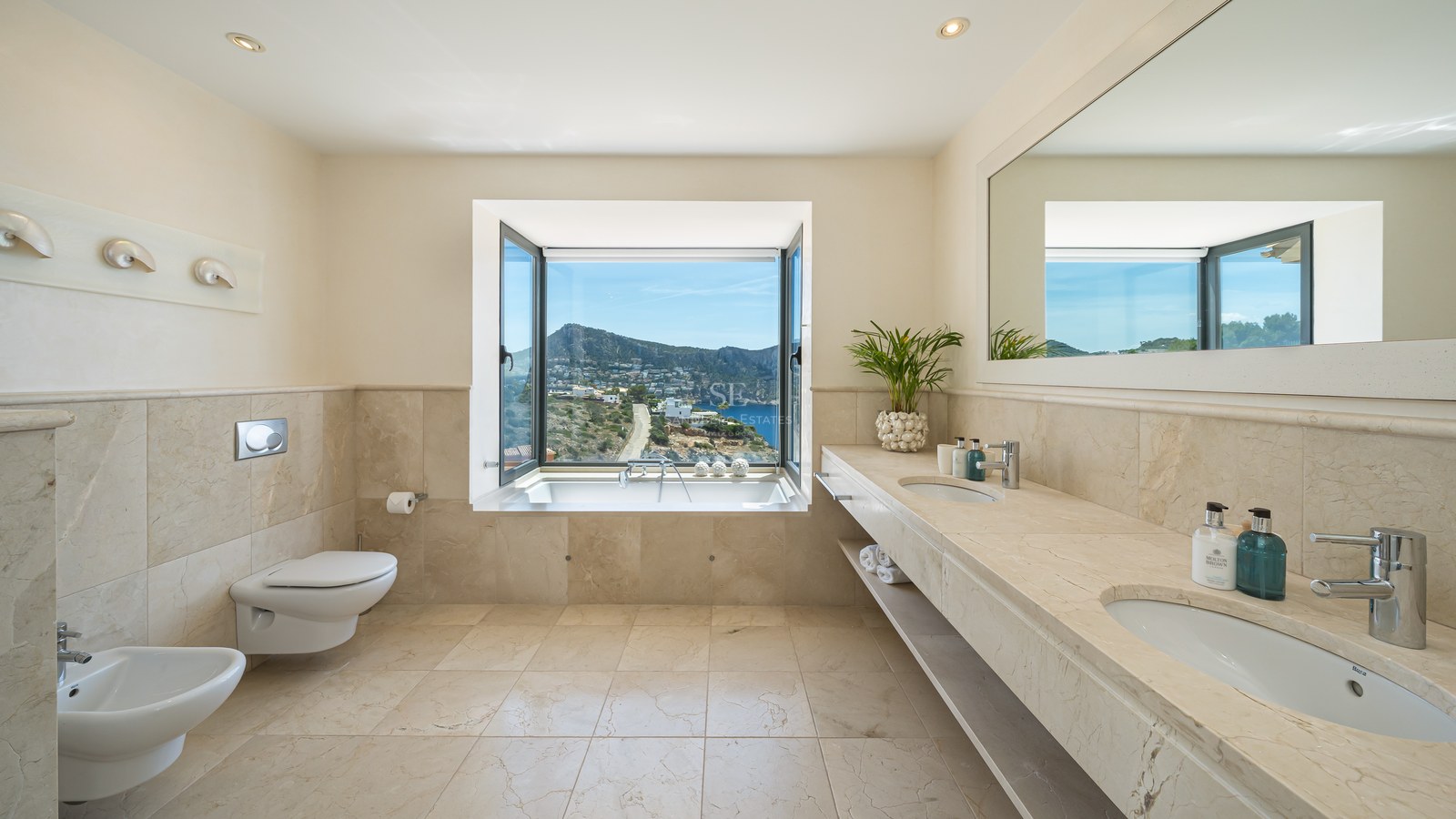 Salle de bain en travertin beige avec double vasque et baignoire face à une fenêtre ouvrant sur la mer et les montagnes.