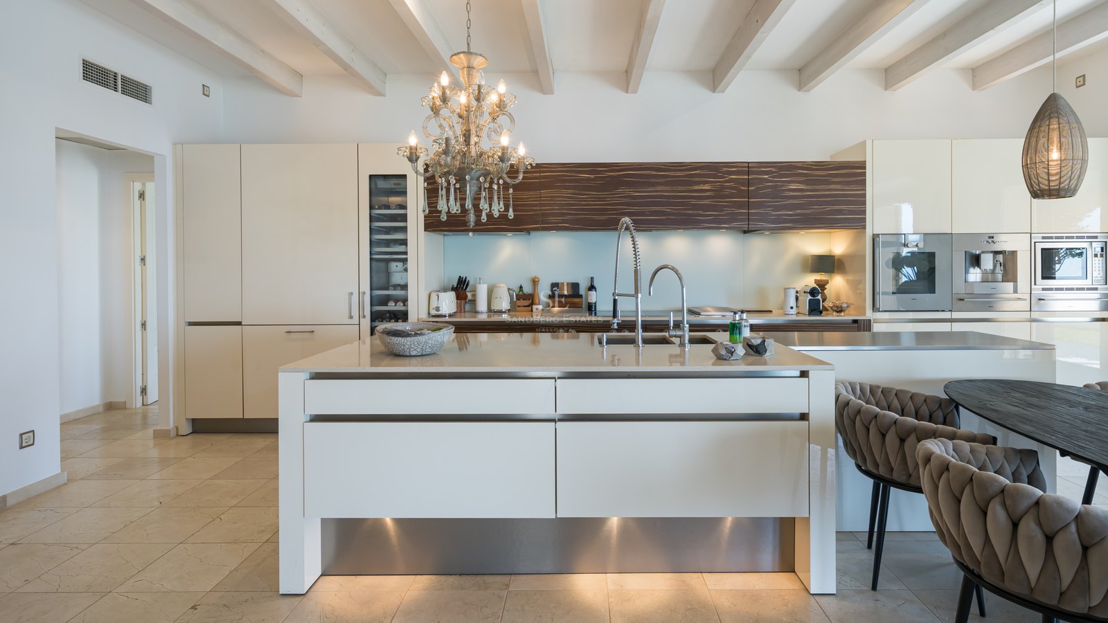 Cuisine moderne blanche avec grand îlot, lustre en cristal et plafonds à poutres apparentes.