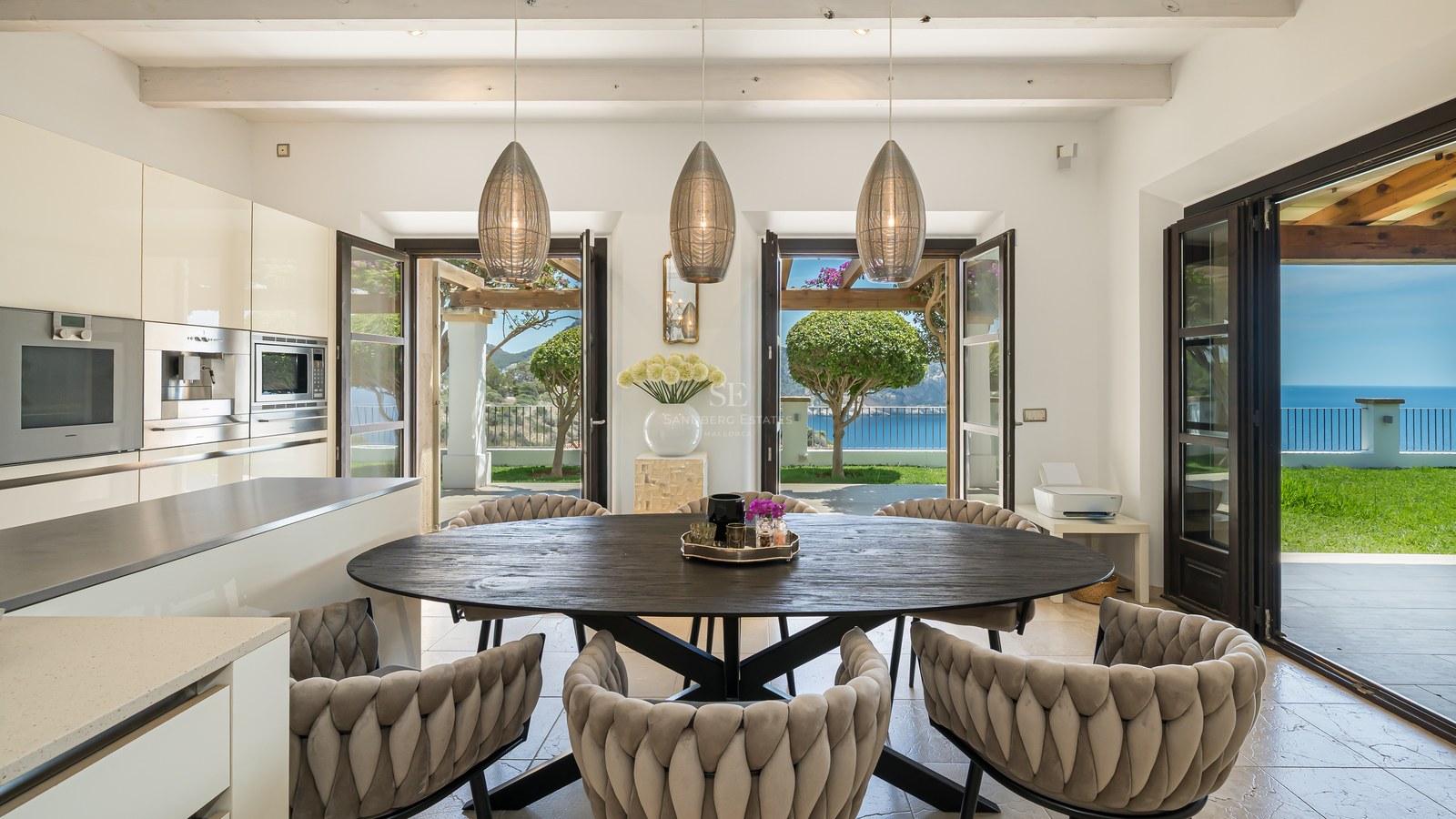 Cuisine moderne avec table ovale en bois sombre, suspensions design et portes vitrées ouvrant sur la mer.