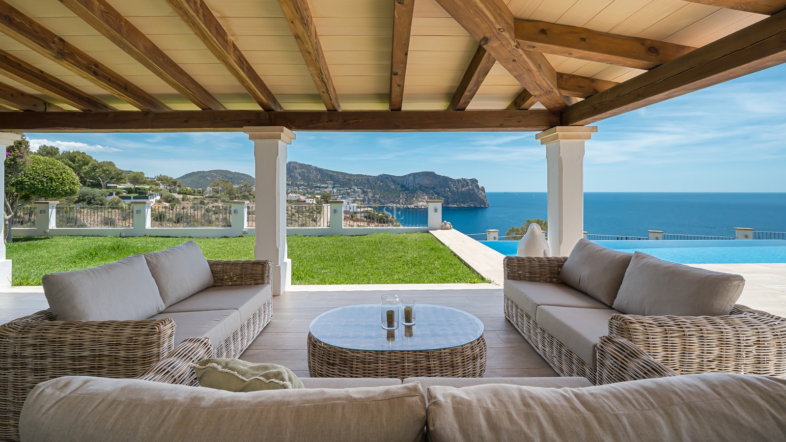 Terrasse couverte avec canapés en osier et poutres en bois donnant sur un jardin, une piscine et la mer.