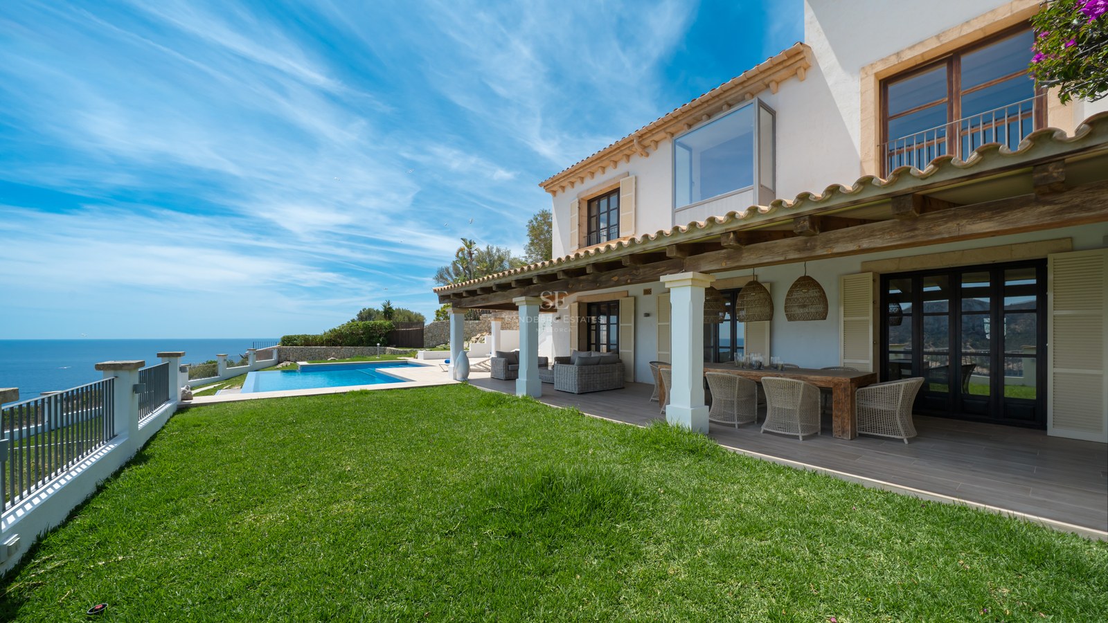 Façade de villa blanche avec terrasse couverte, pelouse et piscine surplombant la mer sous un ciel bleu.
