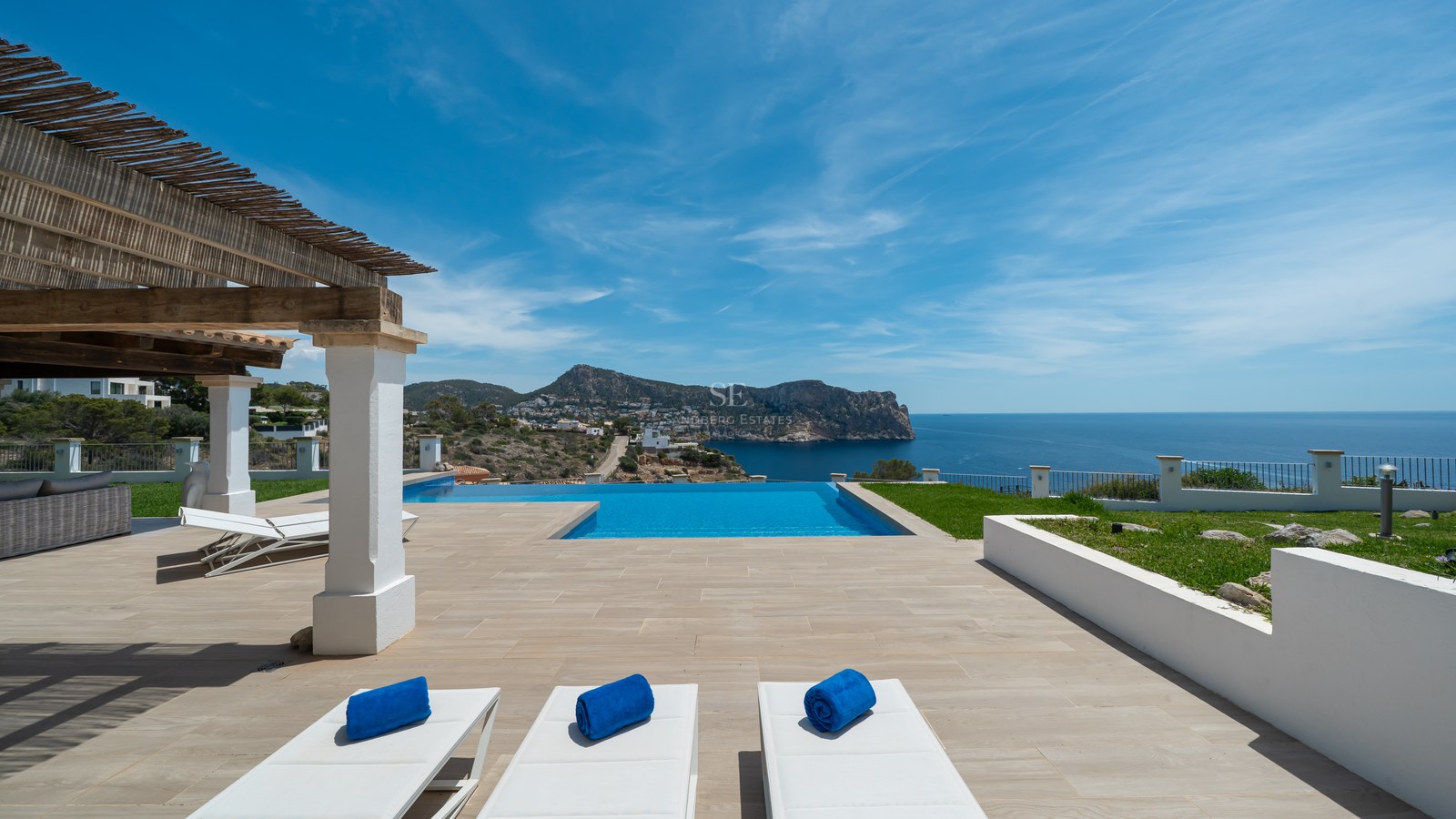 Terrasse de luxe avec piscine à débordement, bains de soleil blancs et pergola en bois surplombant la côte.