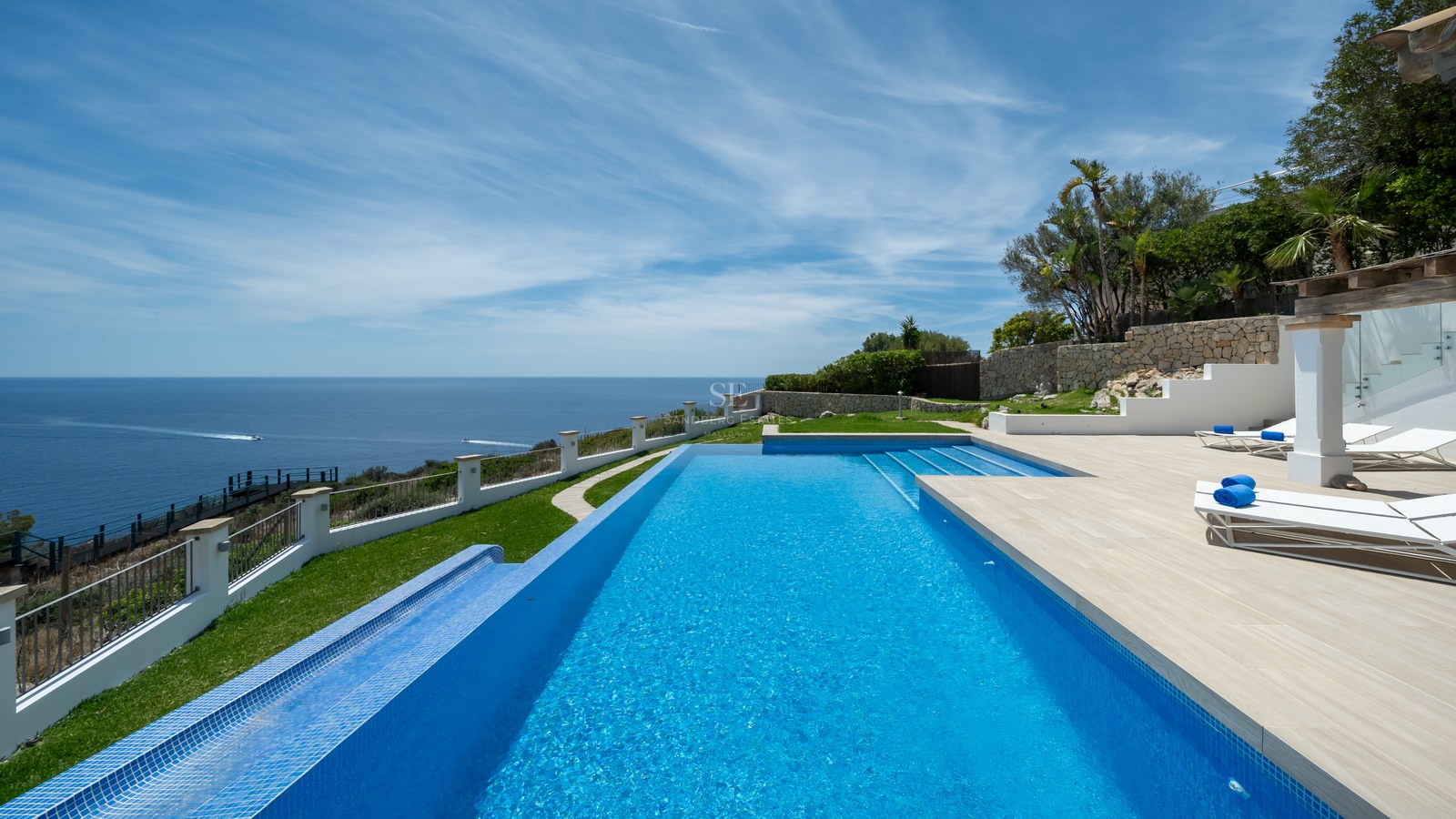 Piscine à débordement bleue surplombant la mer avec transats sur une terrasse en pierre sous un ciel bleu.