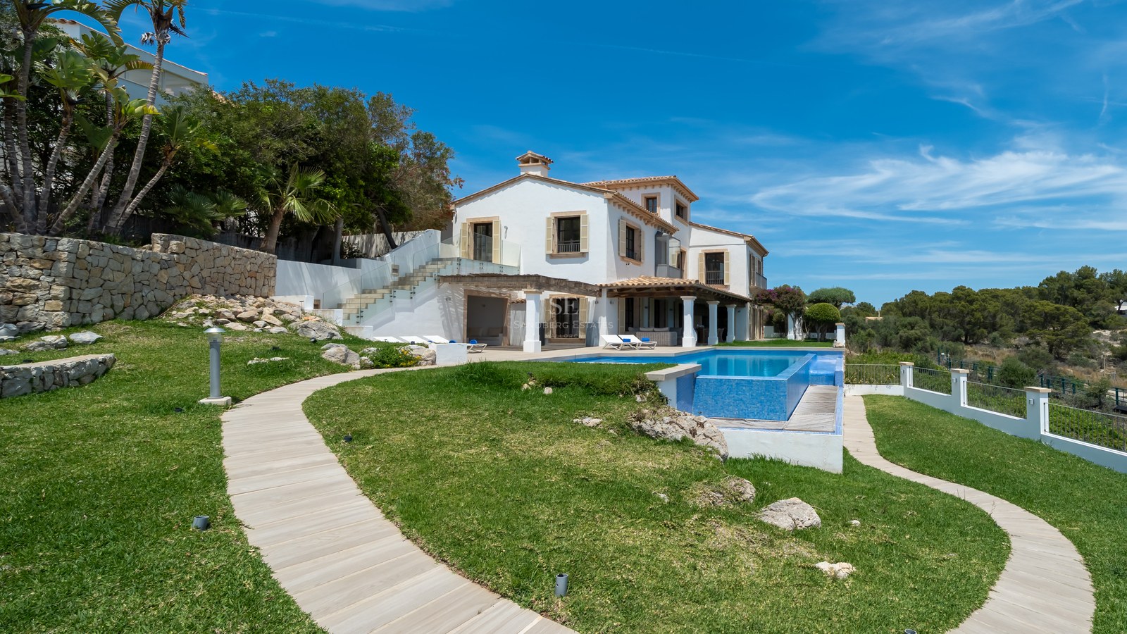 Villa méditerranéenne blanche avec piscine bleue, jardin paysager et chemin en bois sous un ciel bleu.