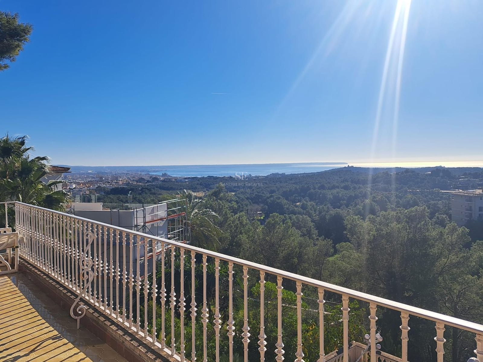 Balcón de forja blanca con vistas a un denso bosque de pinos y al mar Mediterráneo bajo un sol radiante.