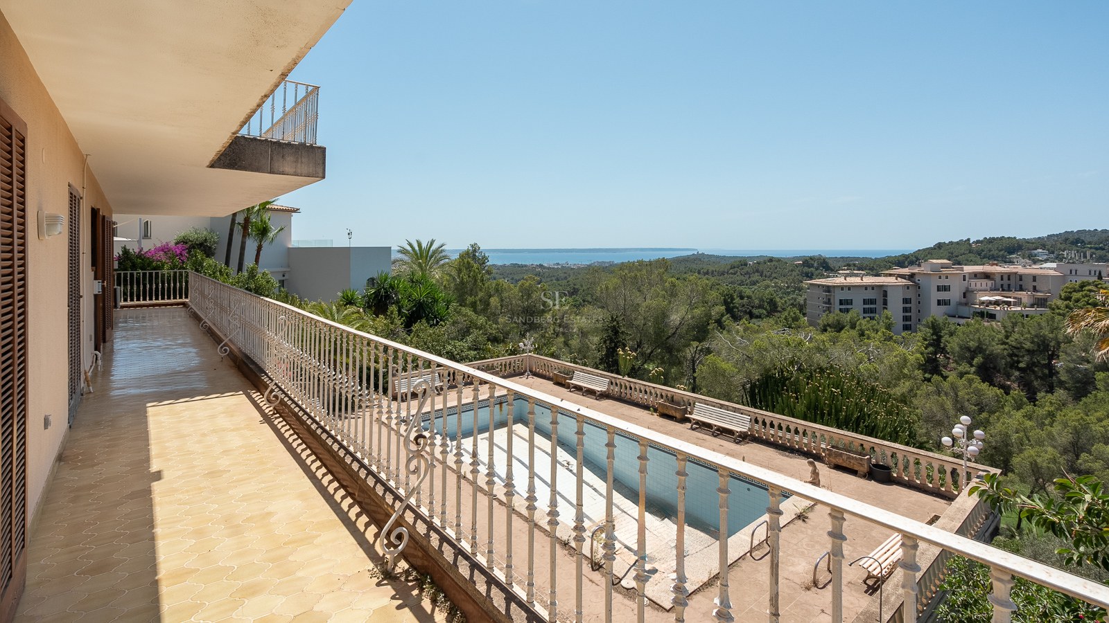 Terraza elevada con barandilla de hierro blanco, vistas a la piscina, pinos y el mar Mediterráneo al fondo.