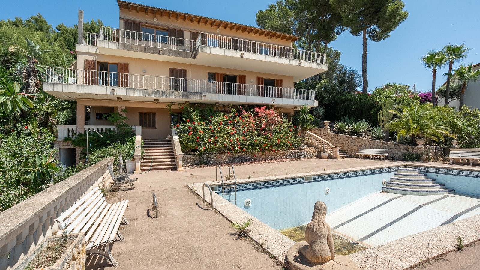 Villa mediterránea de varios niveles con amplias terrazas y una gran piscina vacía rodeada de vegetación exuberante.