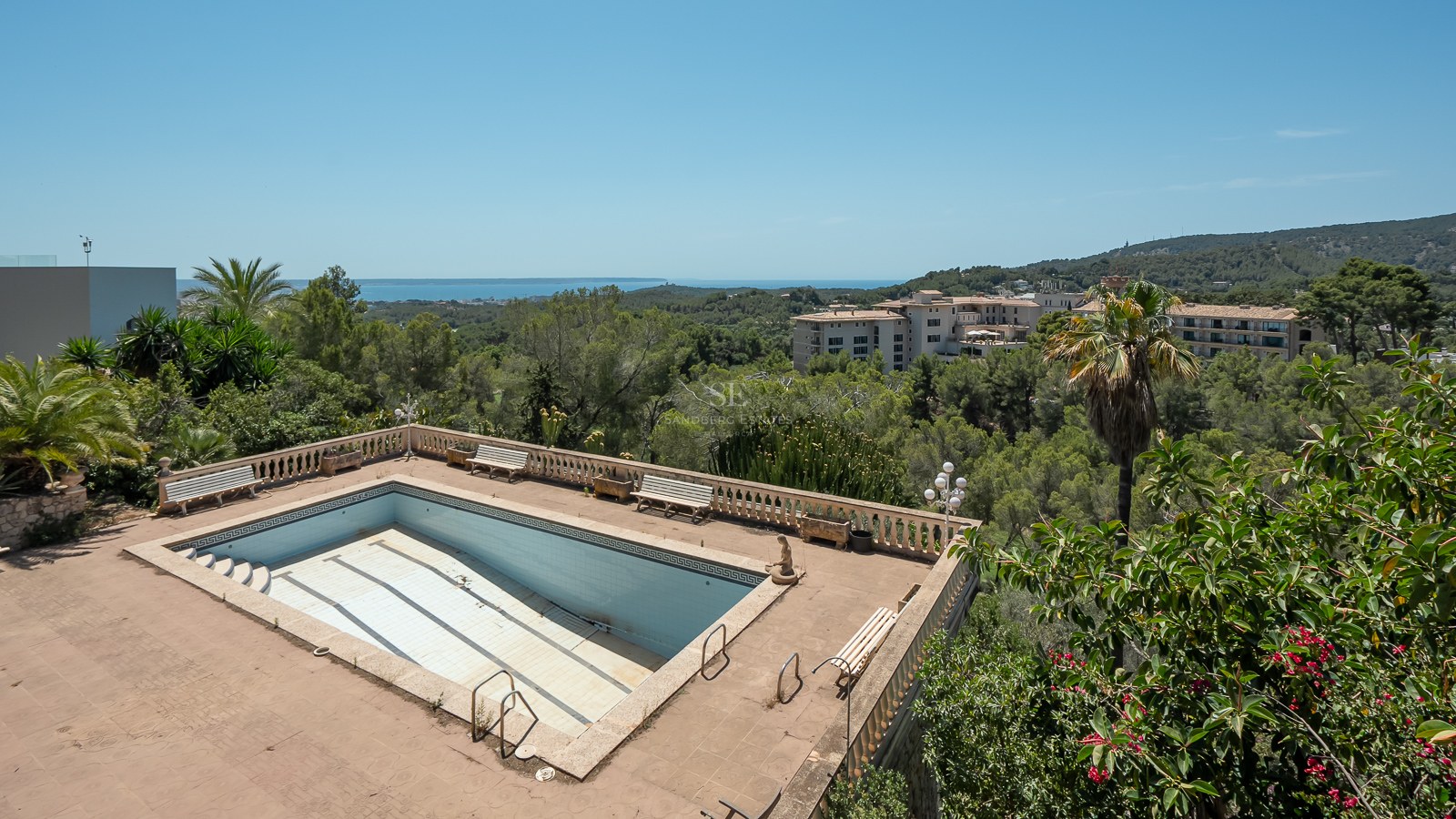 Piscina con balaustrada de piedra con vistas a un bosque frondoso y al mar Mediterráneo al fondo bajo un cielo despejado.