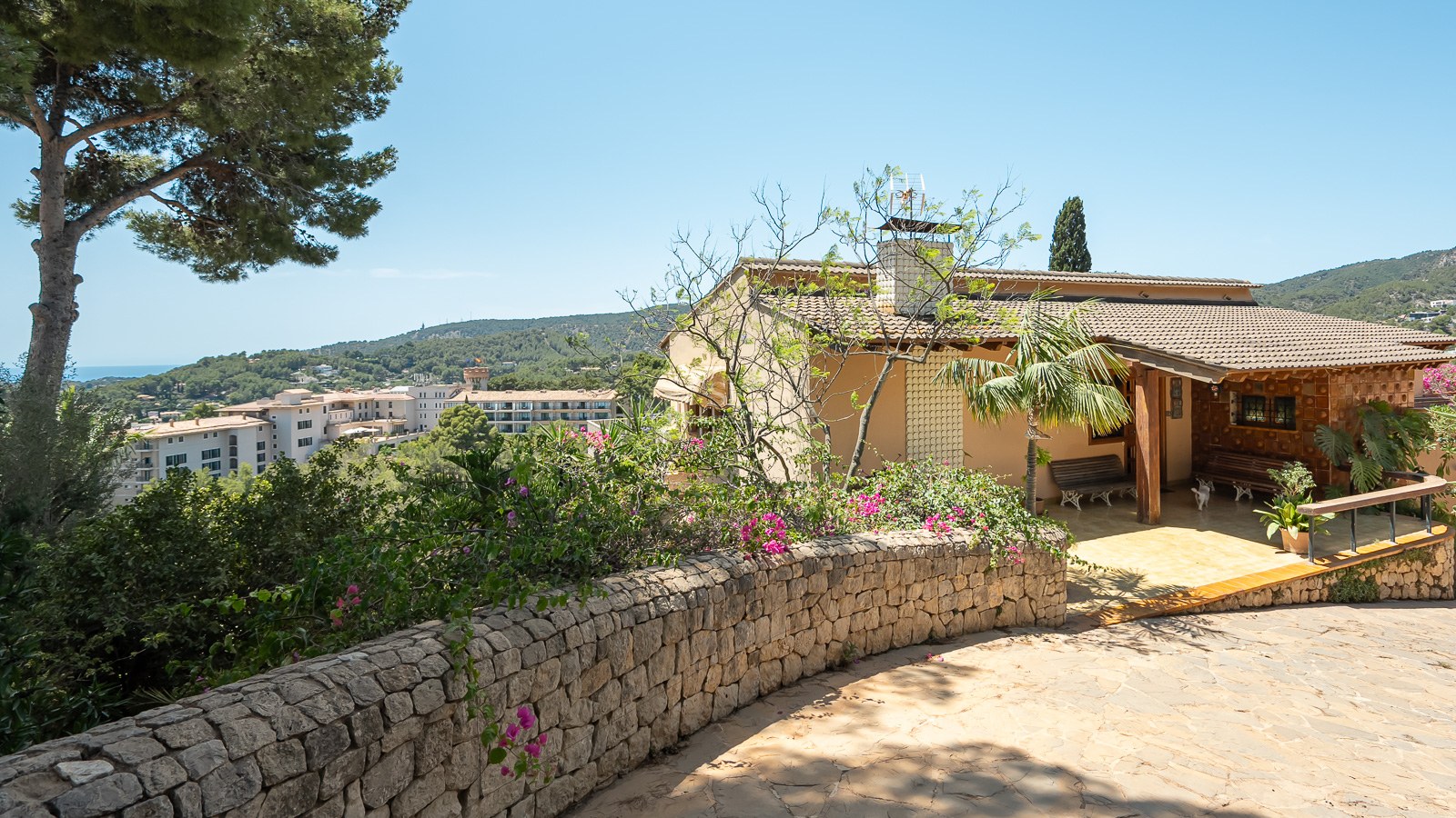 Villa tradicional mediterránea con muro de piedra y porche con vistas a las colinas y el mar al fondo.