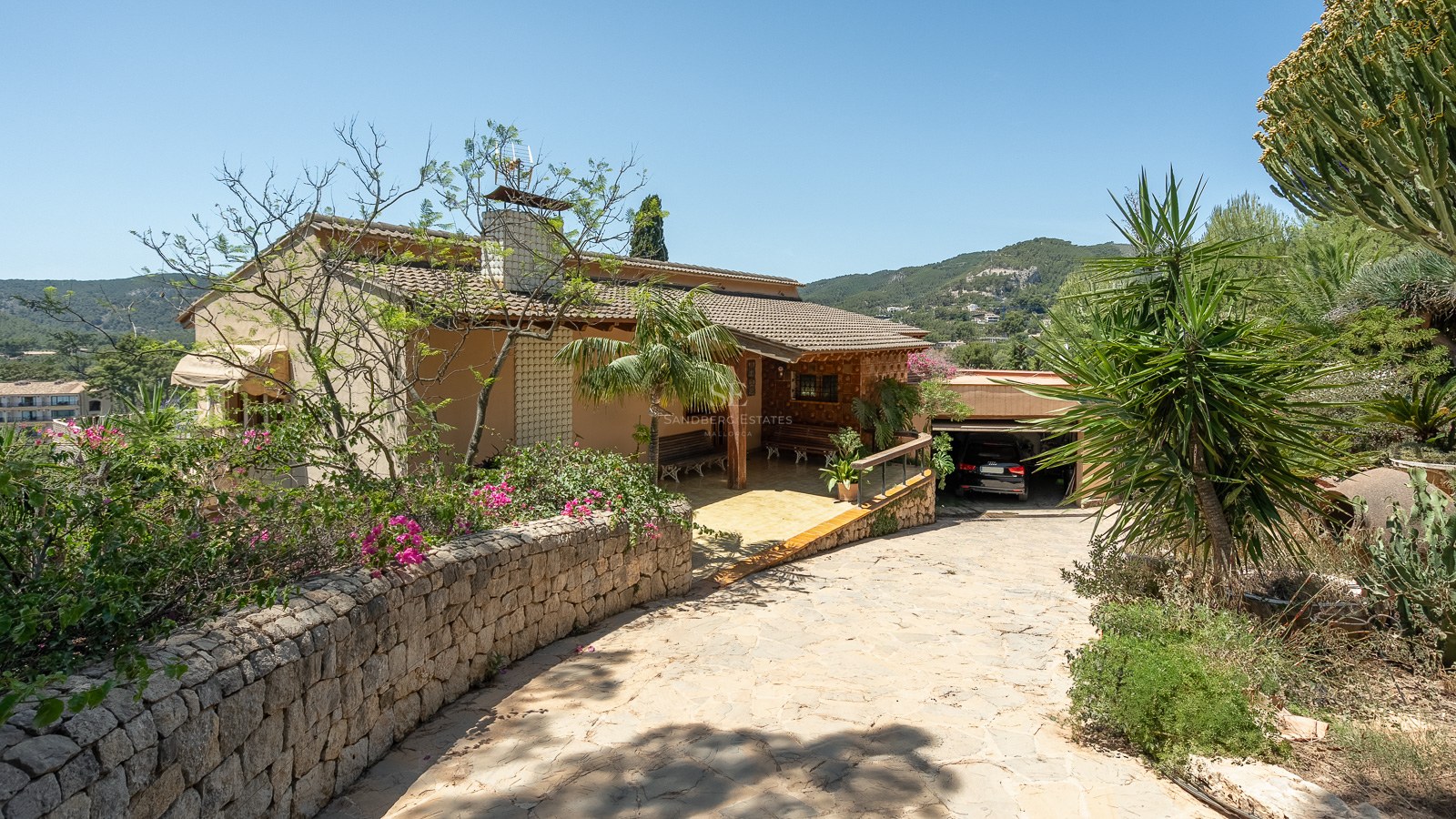 Villa tradicional mediterránea con entrada de piedra, tejado de terracota y garaje rodeada de vegetación exuberante.