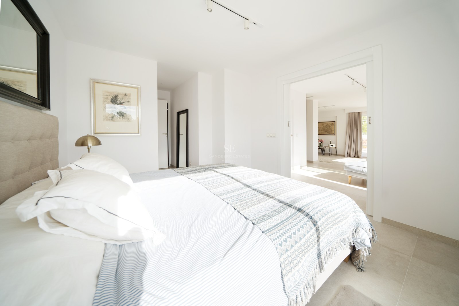Dormitorio principal luminoso con paredes blancas, suelos cerámicos grandes, cabecero acolchado y vistas al salón.