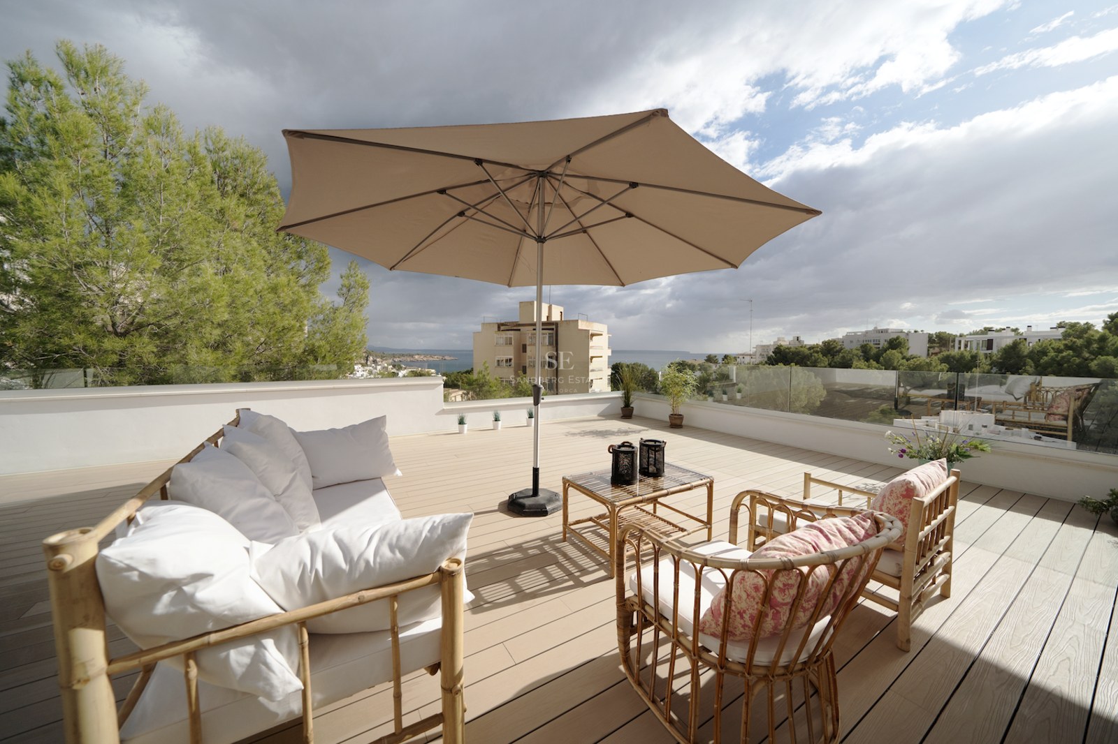 Terraza en azotea con suelo de madera, muebles de bambú, sombrilla beige y barandillas de cristal frente al mar.