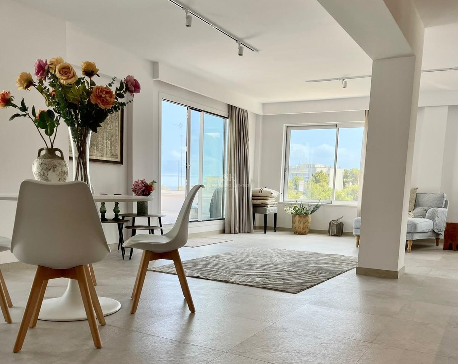 Salón amplio con comedor blanco, alfombra con texturas y grandes ventanales con vistas a la costa y jardines.