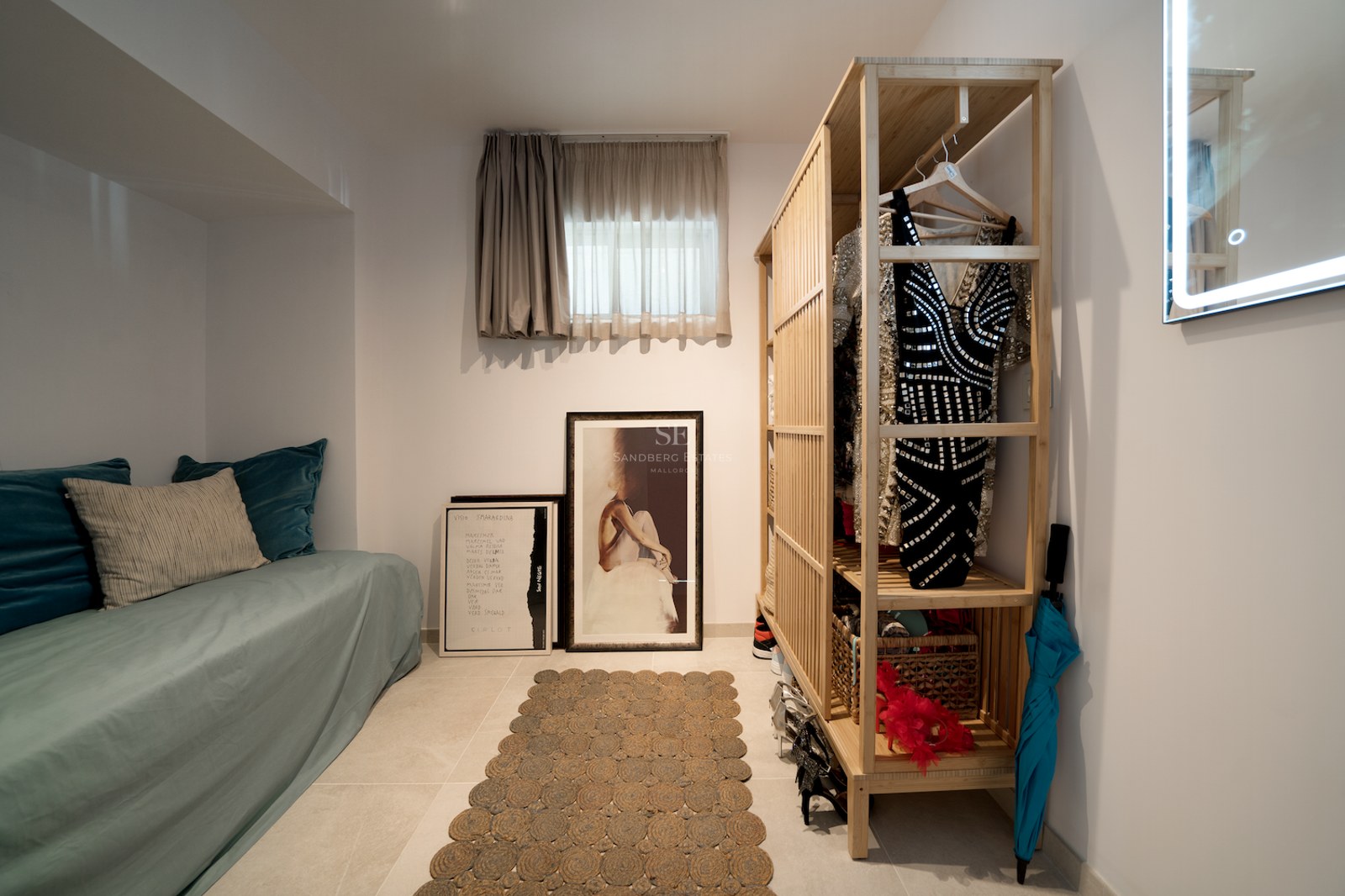 Vestidor moderno con armario de bambú abierto, un diván azul verdoso y cuadros apoyados en el suelo de baldosas.