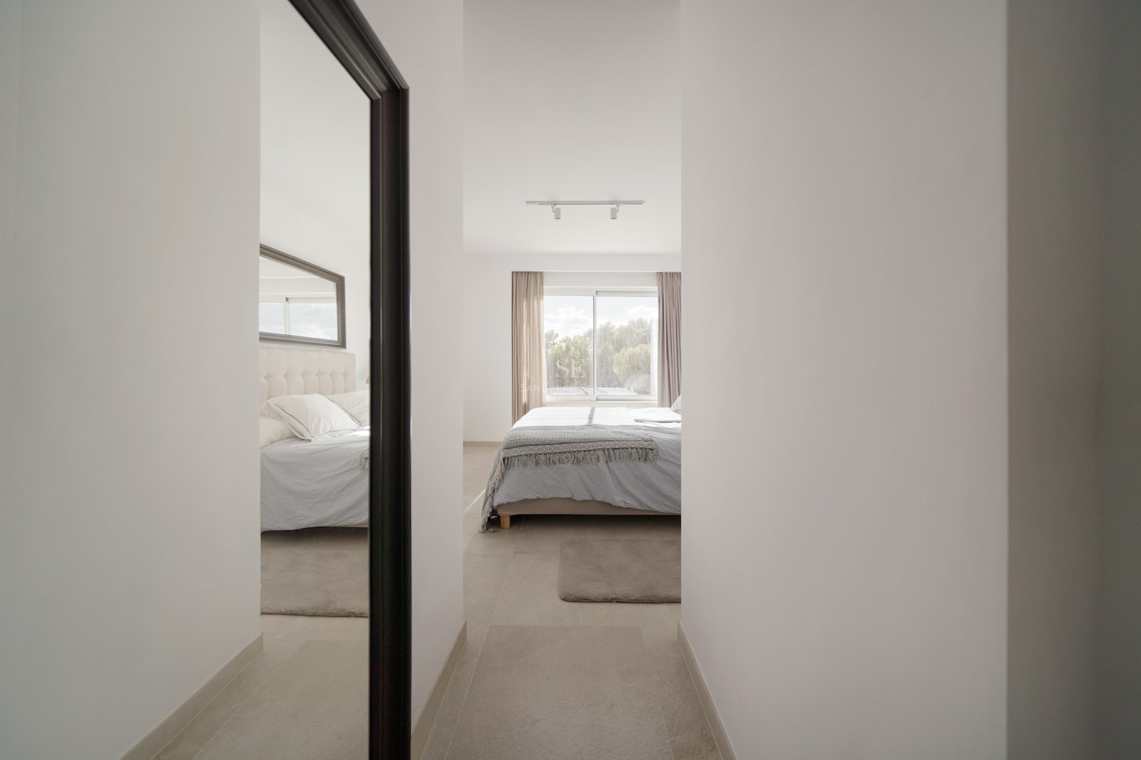 Vista desde un pasillo hacia un dormitorio luminoso con ventanal, textiles neutros y un espejo de cuerpo entero.