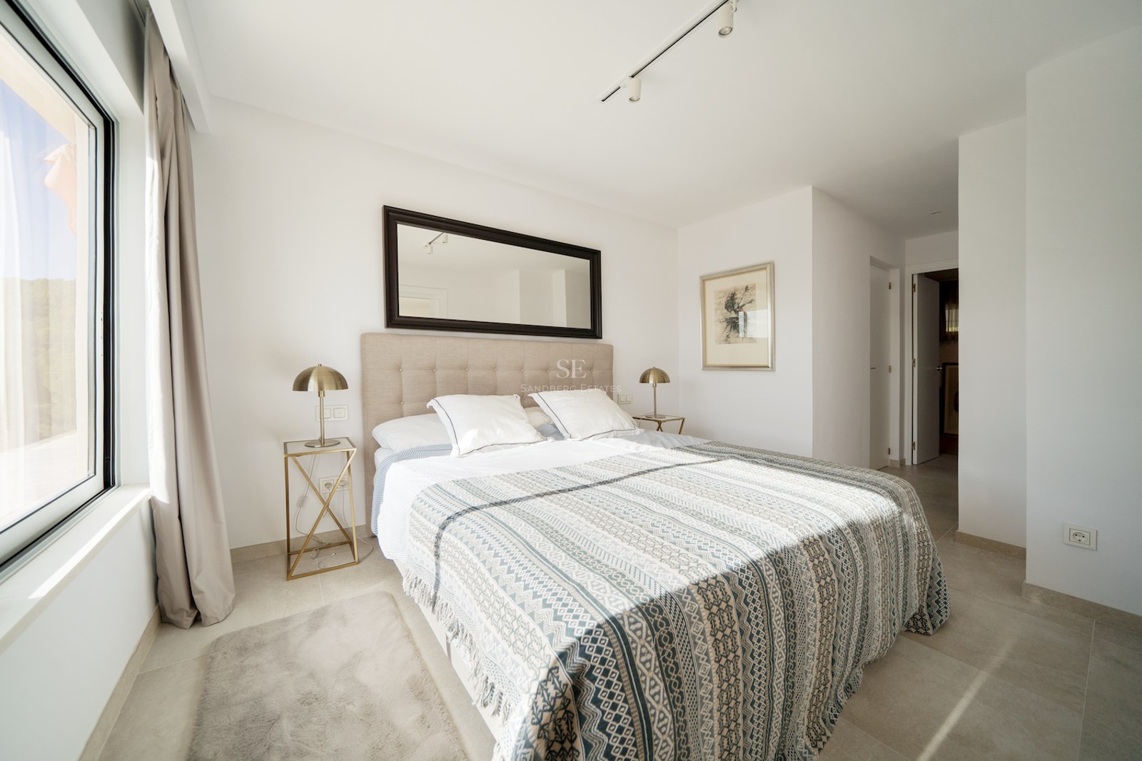 Dormitorio moderno con cama doble, cabecero tapizado, lámparas de latón y un gran espejo bajo luz natural.