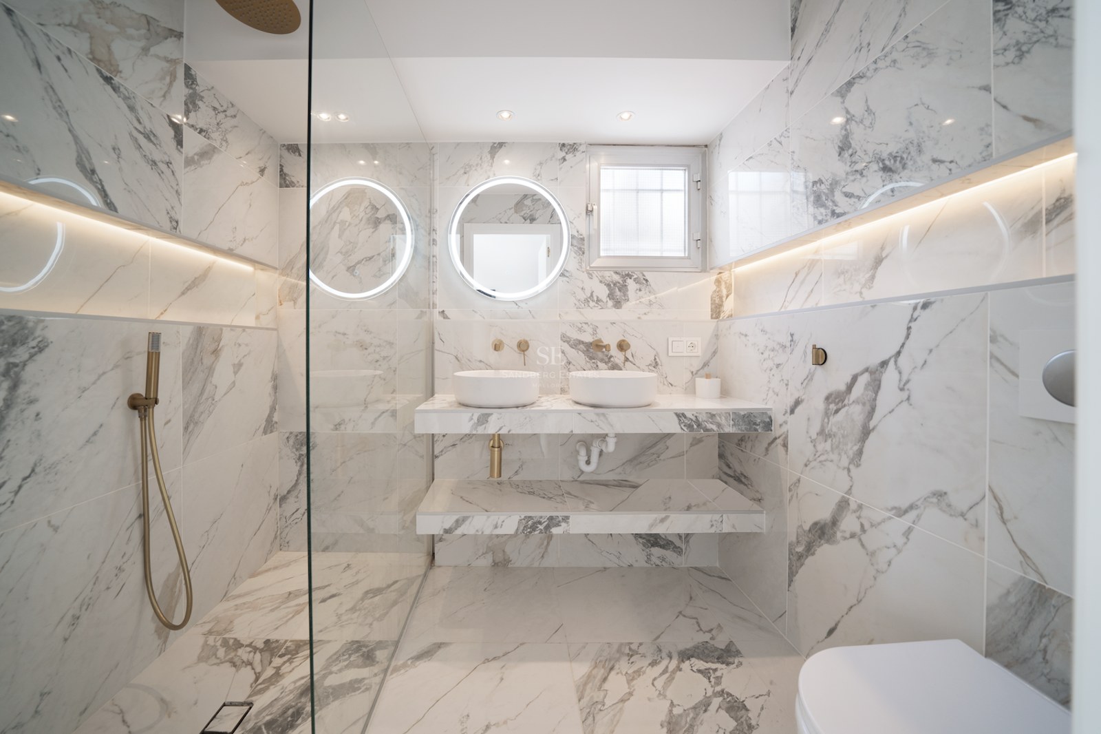 Baño de lujo con paredes de mármol, lavabo doble, espejos circulares retroiluminados y grifería de latón cepillado.