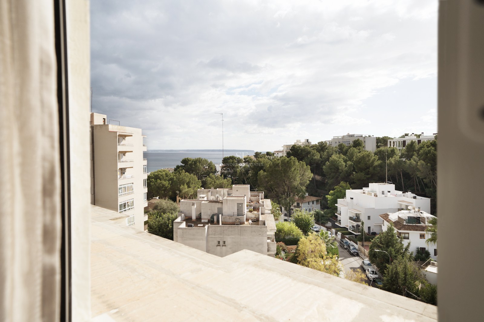 Vista elevada desde una ventana hacia edificios blancos, pinos y el horizonte del mar bajo un cielo nublado.