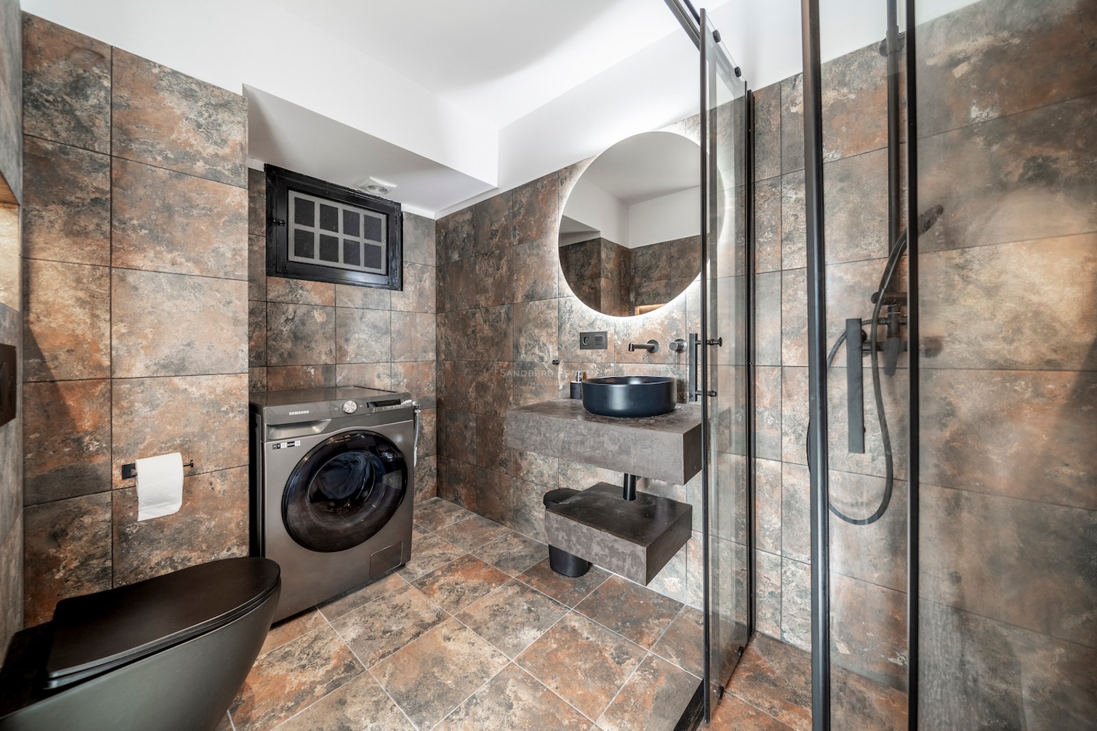 Baño contemporáneo con azulejos efecto piedra, lavabo suspendido negro, espejo retroiluminado y ducha de cristal.