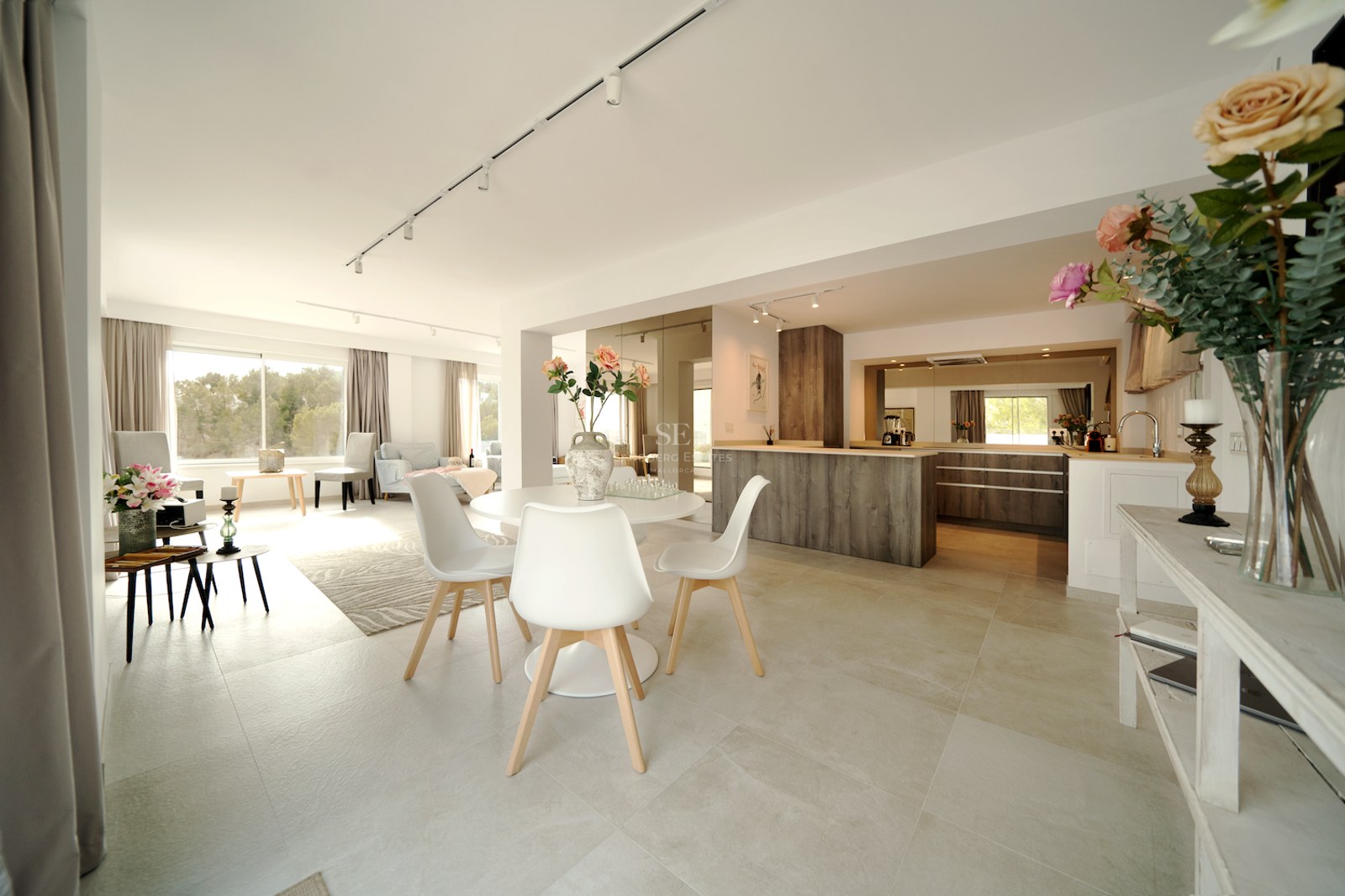 Amplia sala de planta abierta con mesa de comedor blanca, suelos de porcelánico y cocina con acabados en madera.